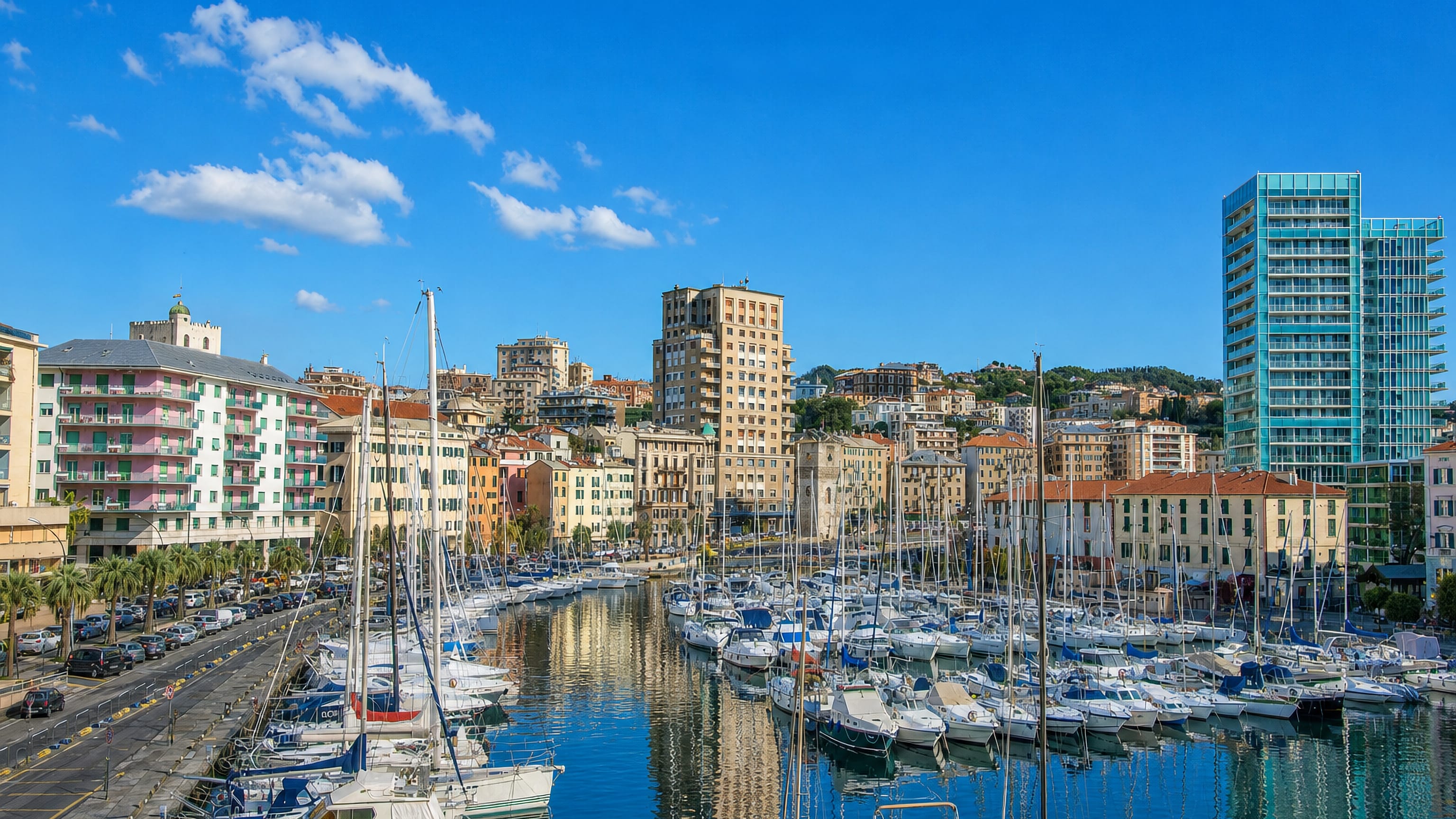 Sailboats moored in Savona's marina below pastel apartment blocks and a modern glass tower on the Ligurian coast.