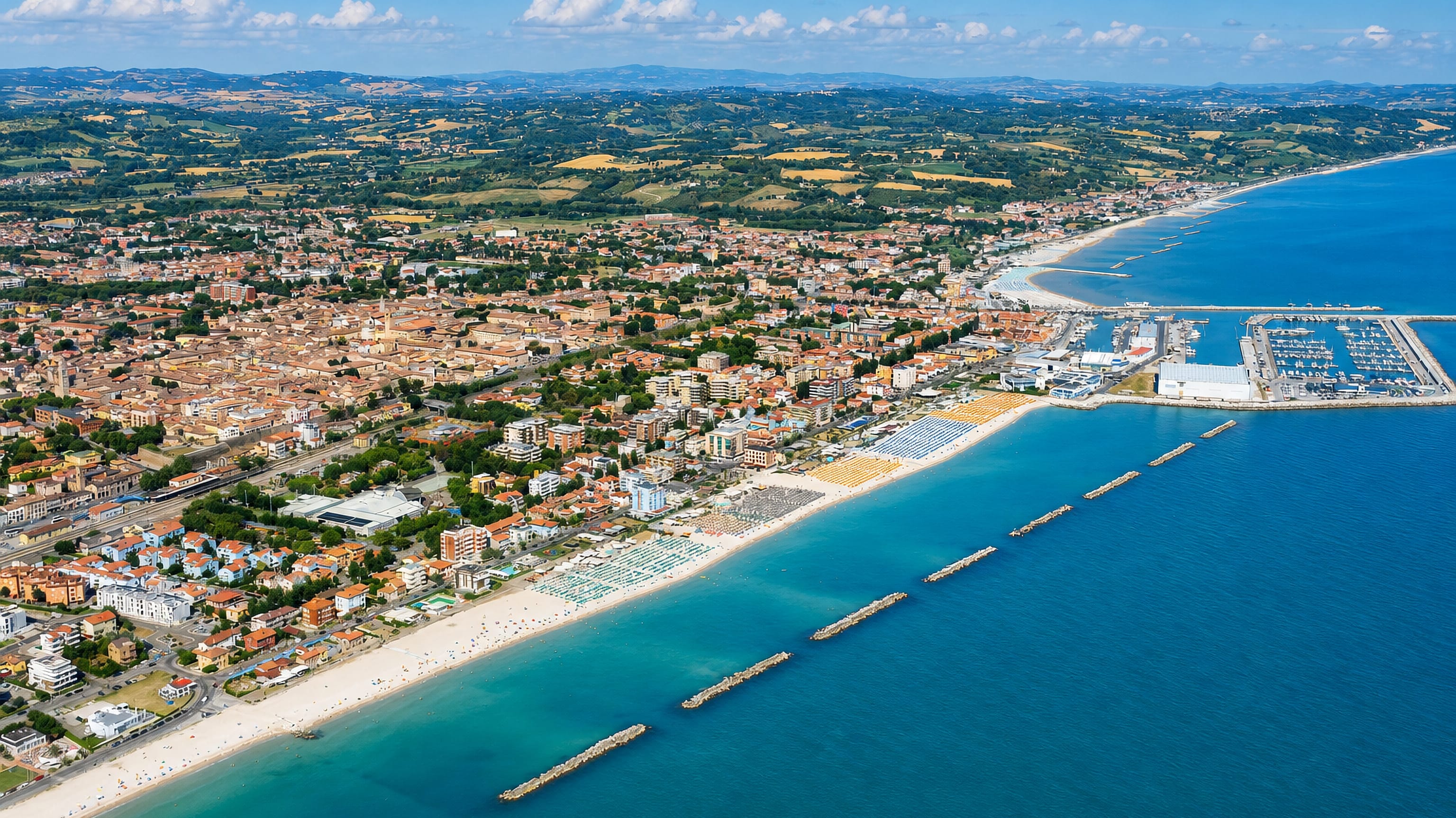 Aerial view of Fano's Adriatic beachfront, breakwaters and marina with the Marche hills behind.