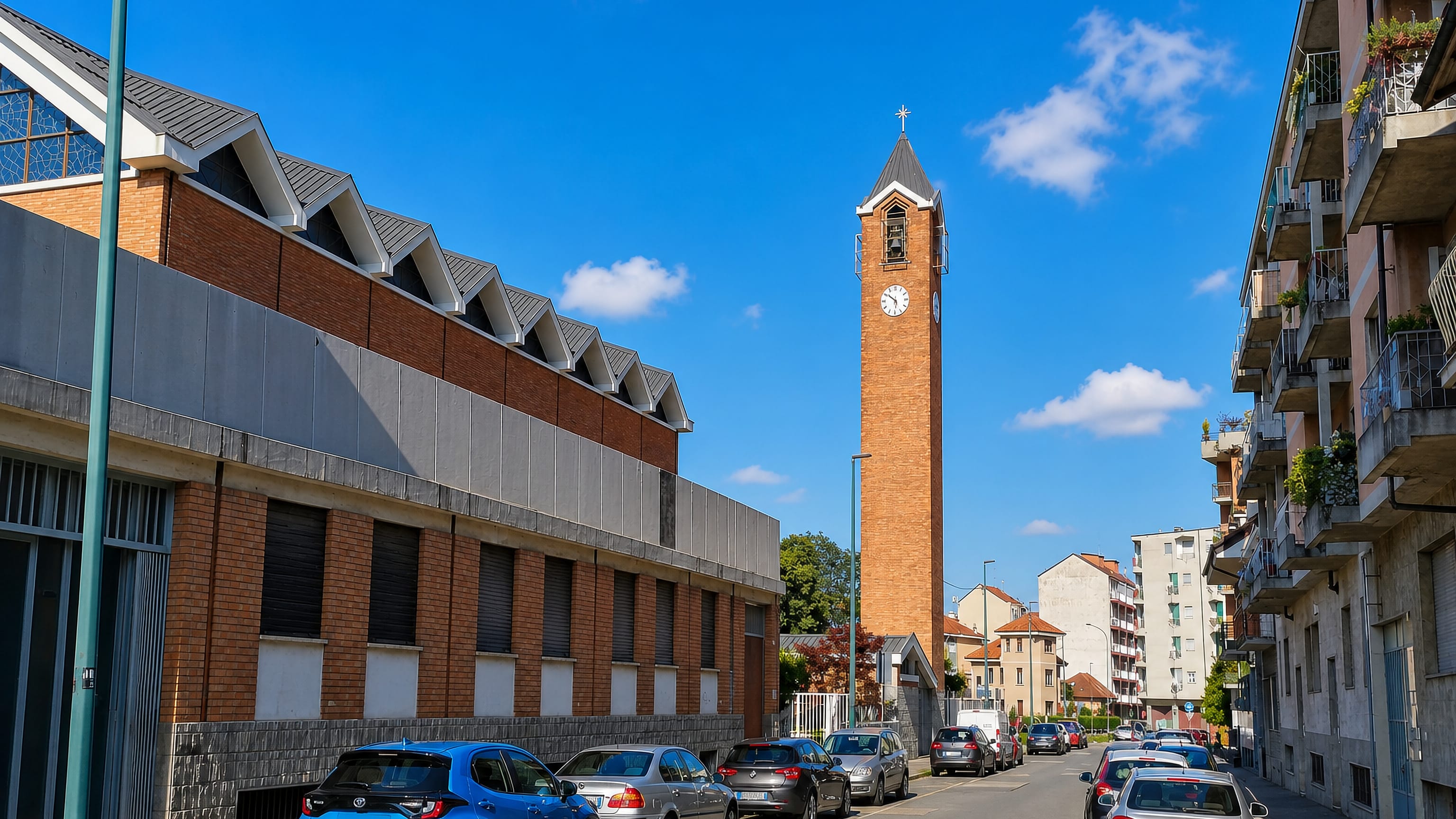 Sunlit street in Parella, Turin, lined with apartment blocks and a tall brick church campanile with a clock.
