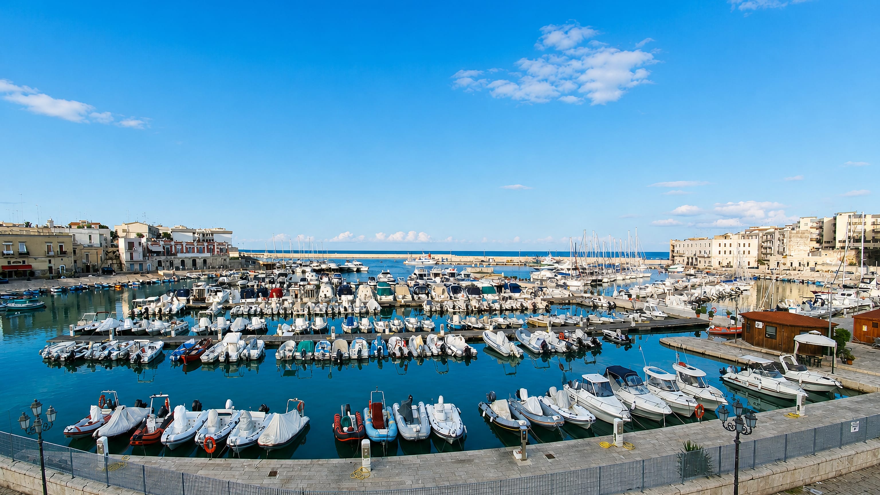 Boats moored in Bisceglie's small harbour basin, ringed by a stone quay and Puglian townhouses.