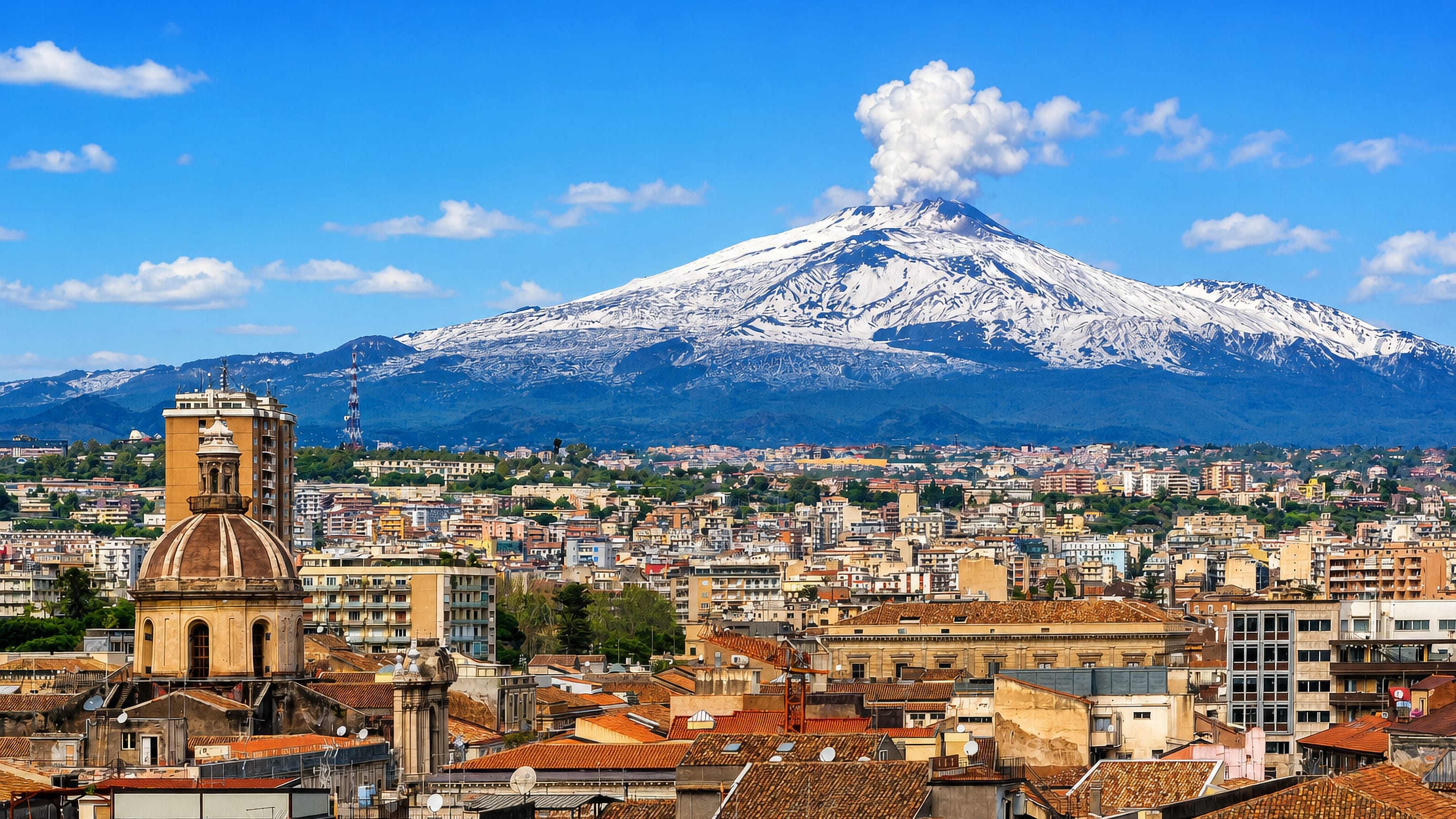 Catania's terracotta rooftops and baroque church dome beneath snow-capped Mount Etna venting a steam plume