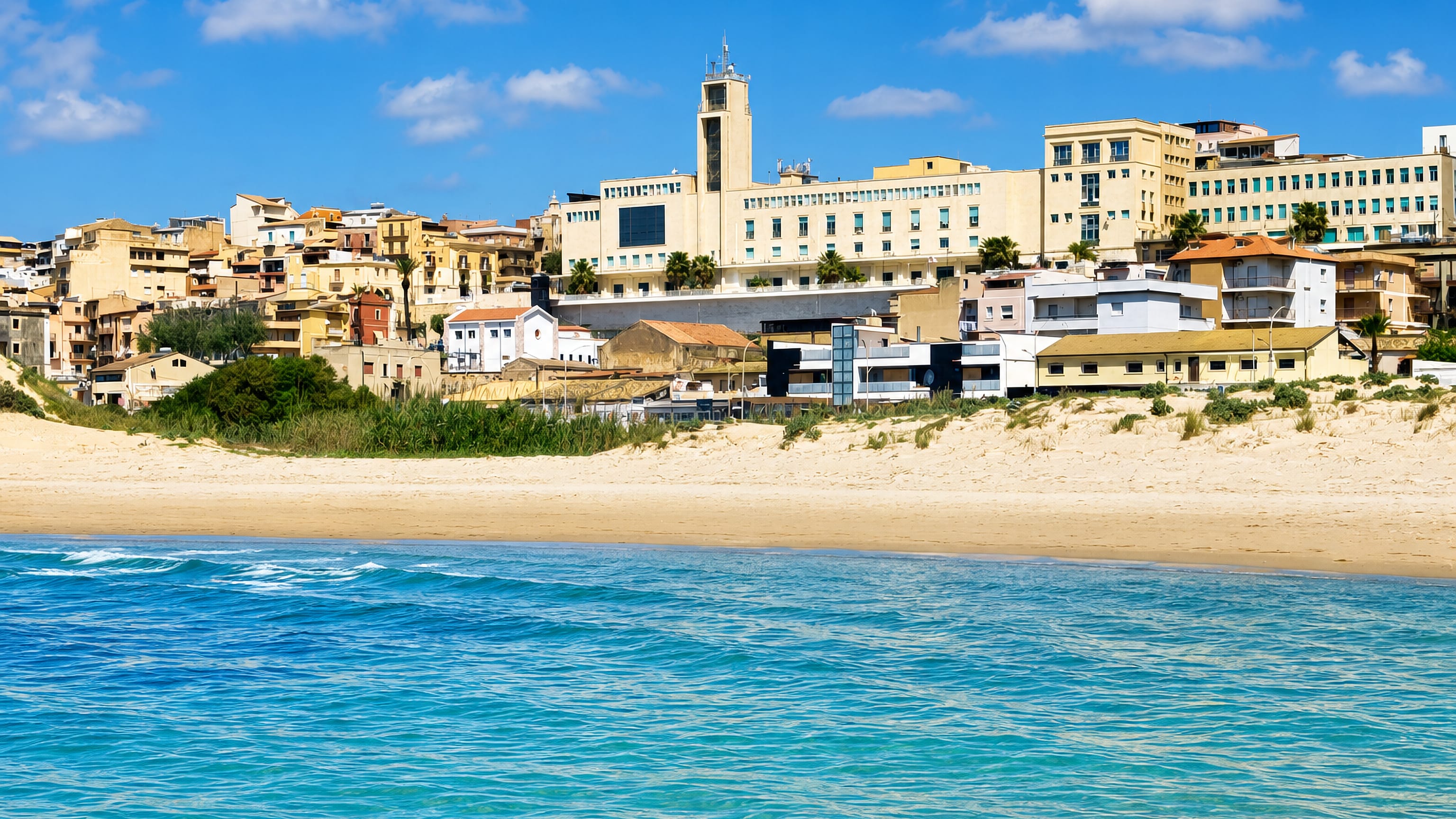 Sandy beach and turquoise sea in front of Gela's hillside houses and pale seafront buildings, Sicily.