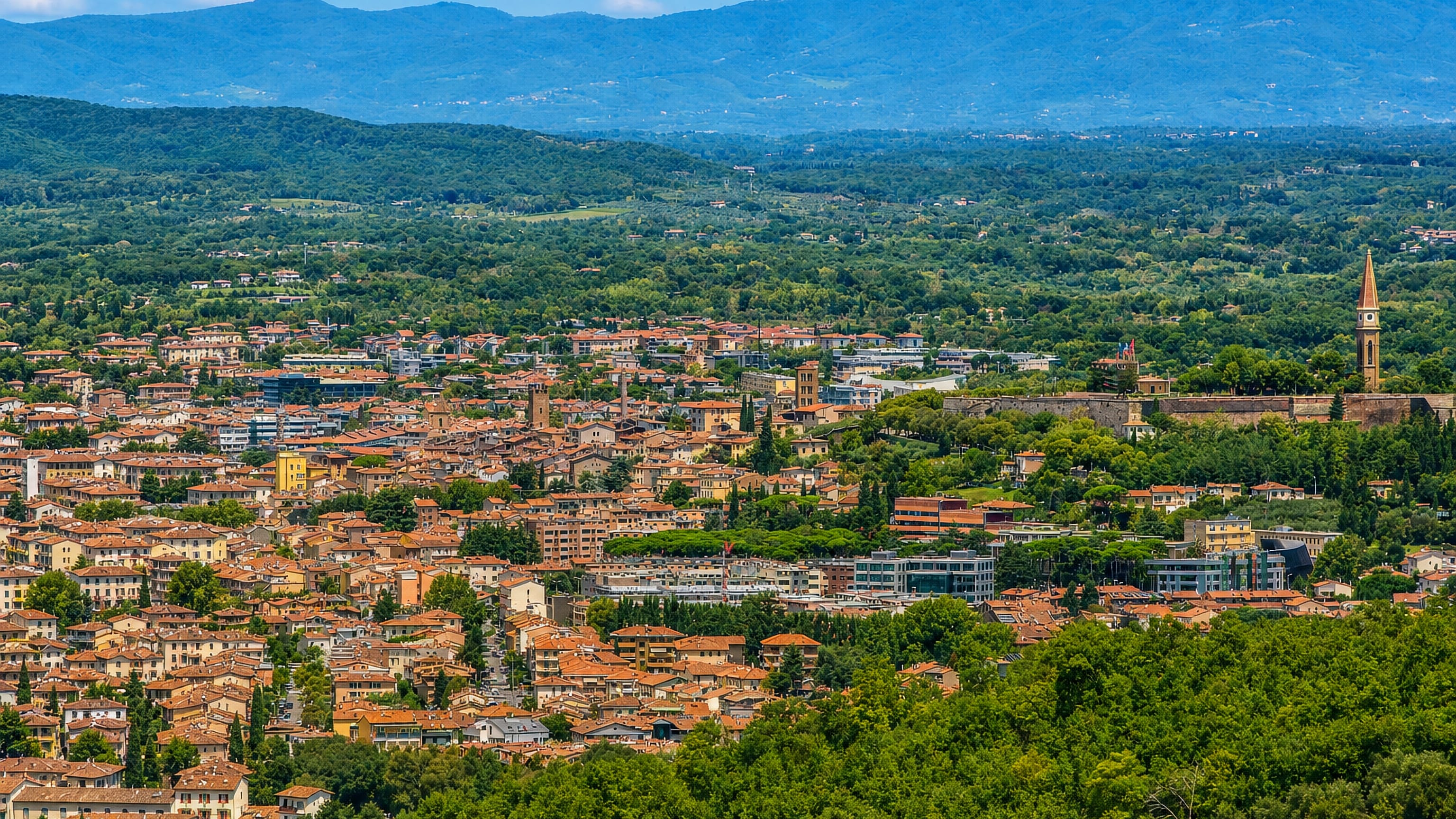 Arezzo's terracotta rooftops and hilltop bell tower below the wooded Tuscan hills under a blue sky.