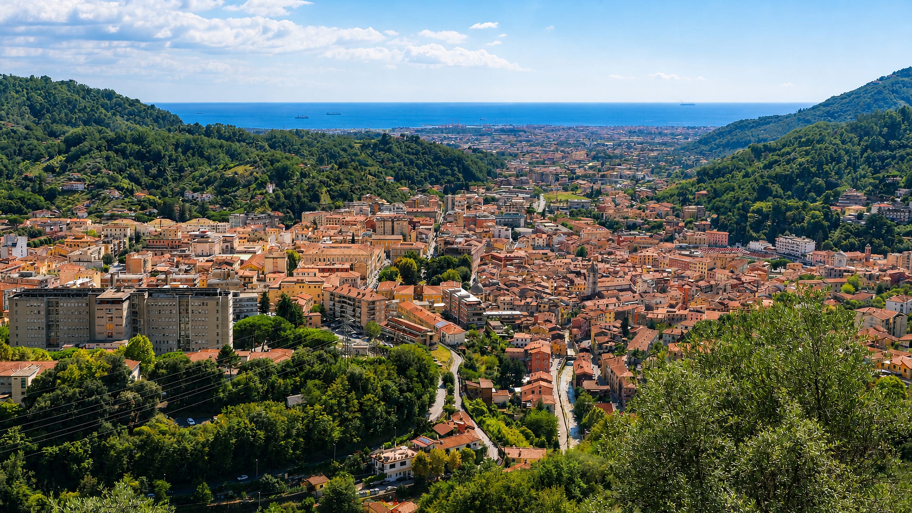 Carrara's terracotta rooftops fill a green Apuan valley opening to the Ligurian Sea and its port on the horizon.