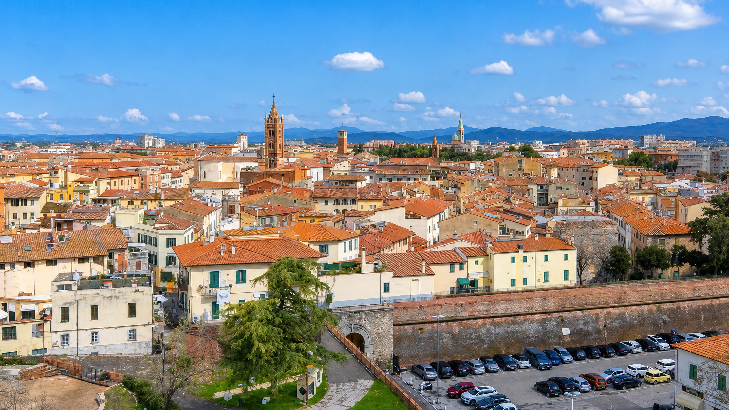Grosseto's terracotta rooftops and bell tower seen over the brick Medici walls, with a car park below