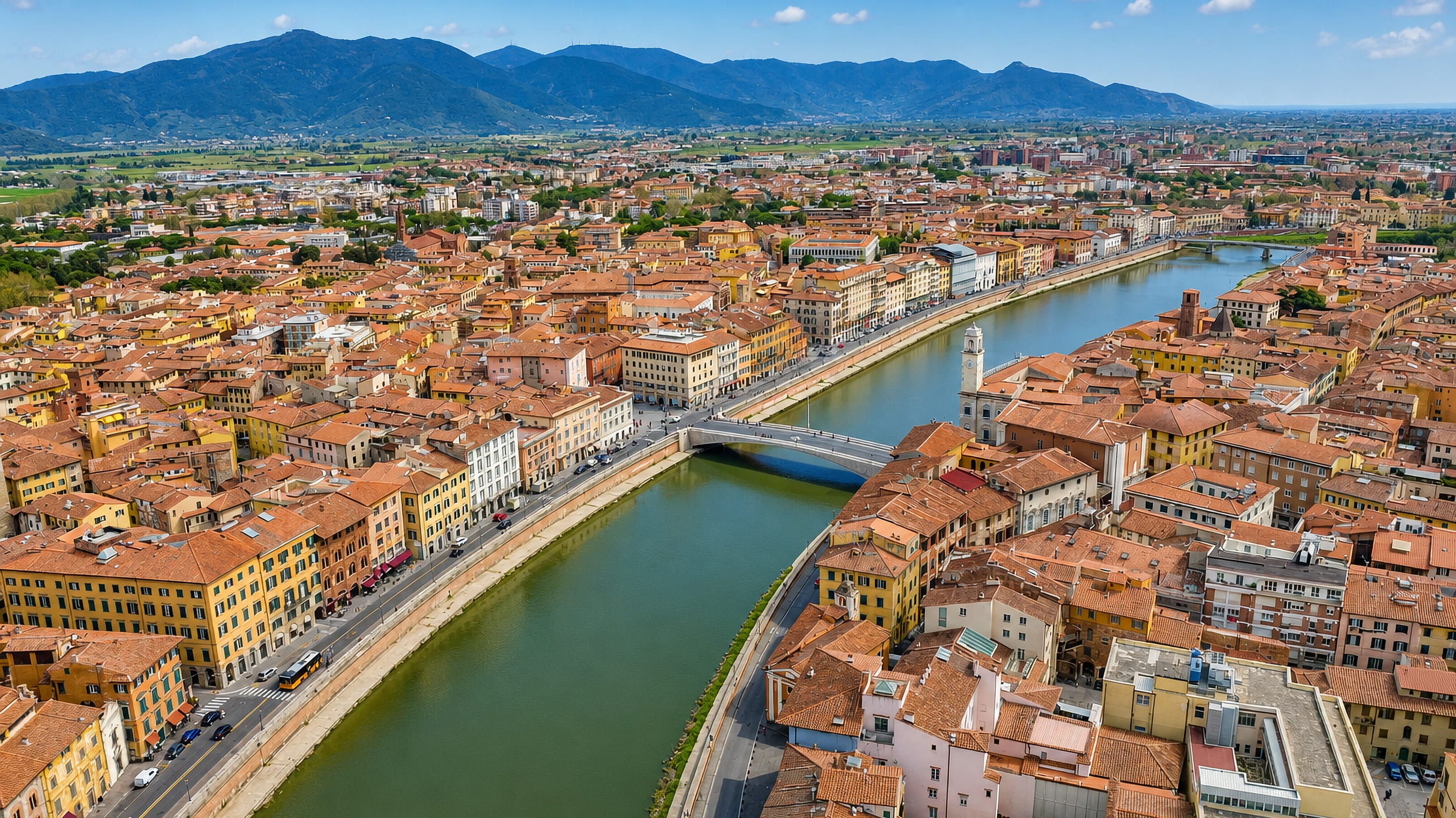 Aerial view over Pisa's Arno river, lined with pastel lungarni palazzi and terracotta roofs below the Monti Pisani