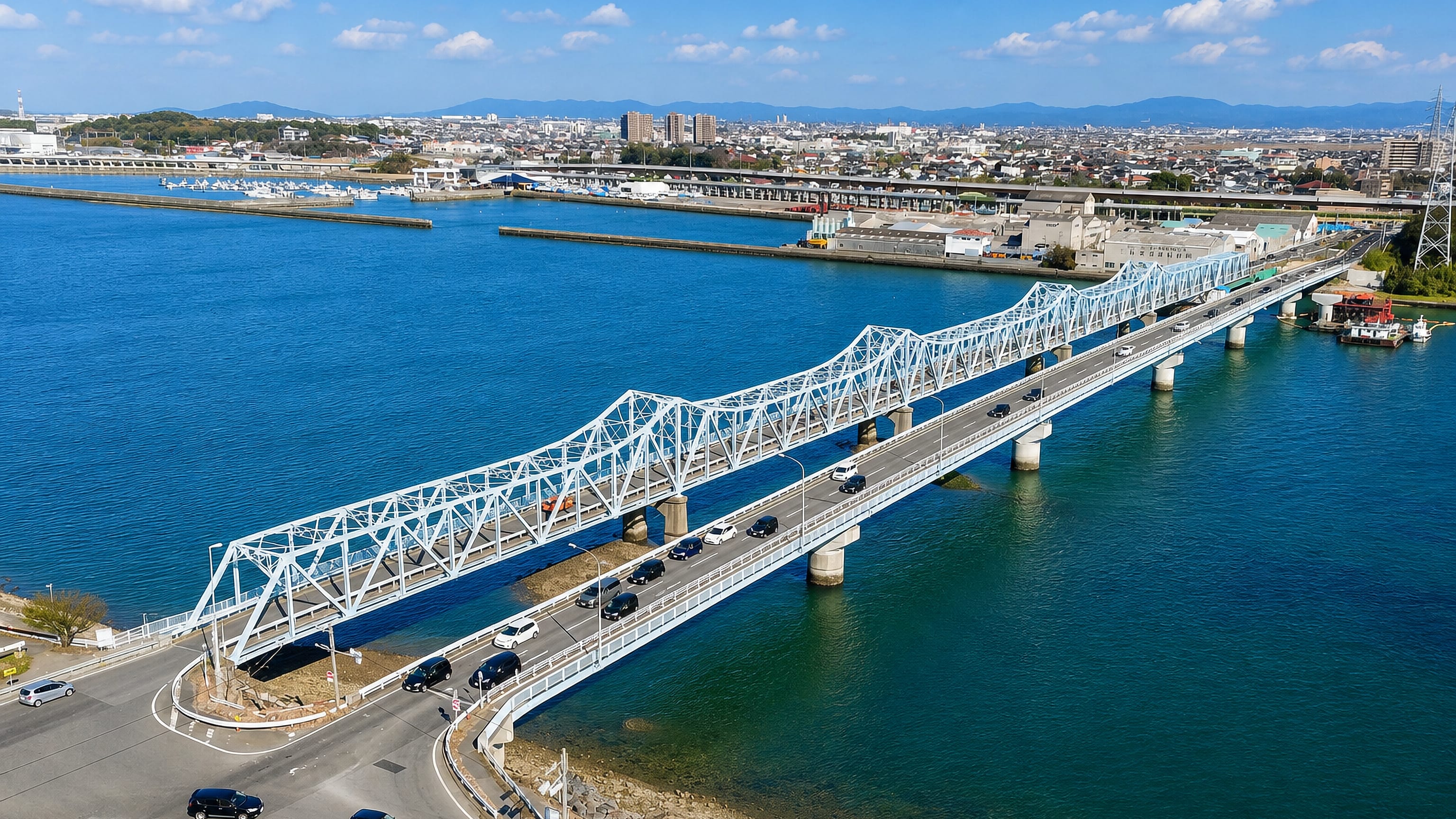 Steel truss road bridge crossing the bay at Handa, Japan, with the town and wooded hills beyond.