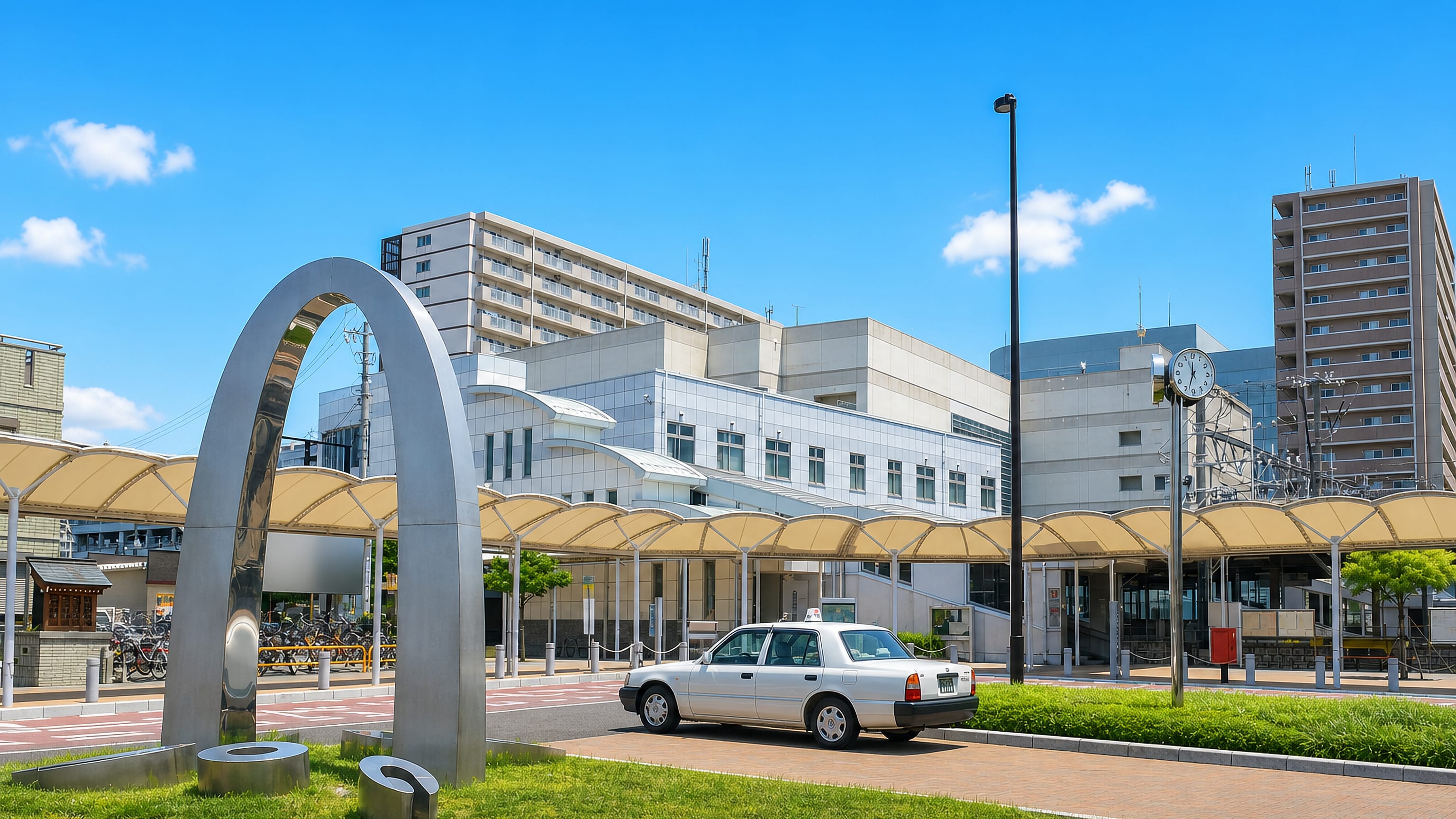 Taxi waiting under the canopied station plaza beside a steel arch sculpture in Kitanagoya, Aichi.