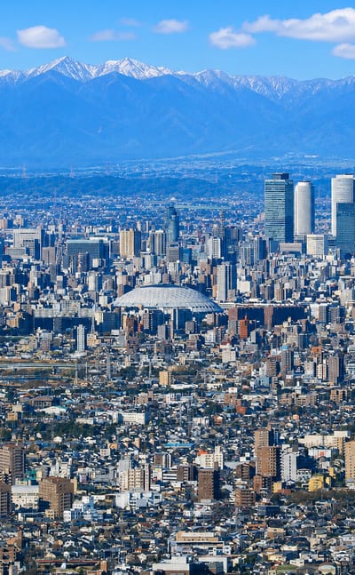 Dense Nagoya cityscape with the JR Central Towers and Nagoya Dome below snow-capped mountains.