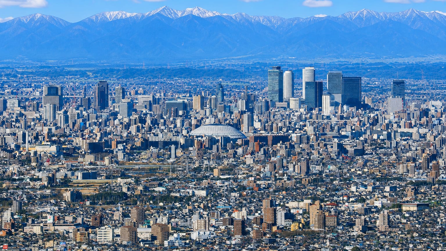 Dense Nagoya cityscape with the JR Central Towers and Nagoya Dome below snow-capped mountains.