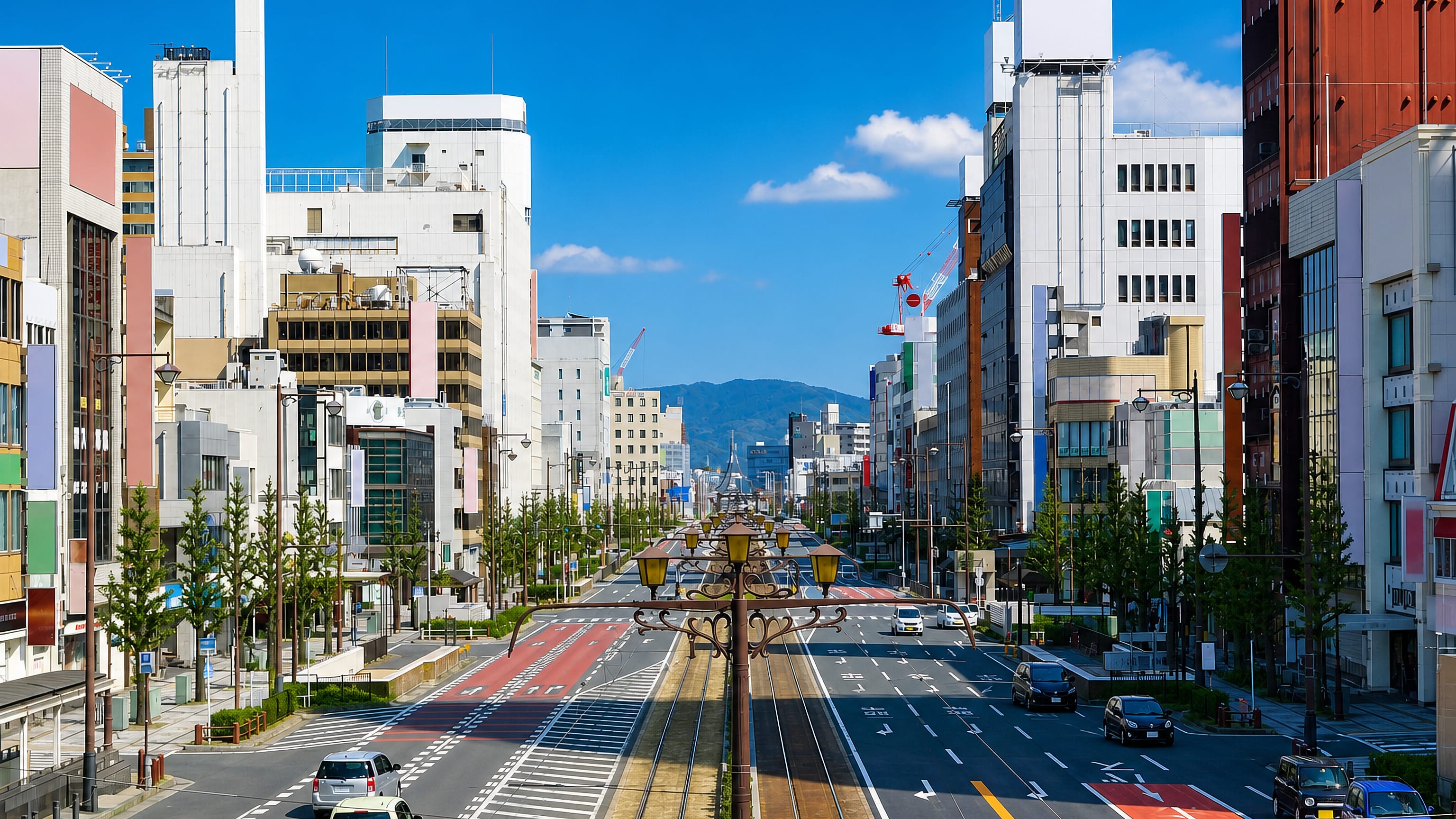 Wide tram-lined boulevard through central Toyohashi, Japan, with mid-rise blocks and hills beyond.