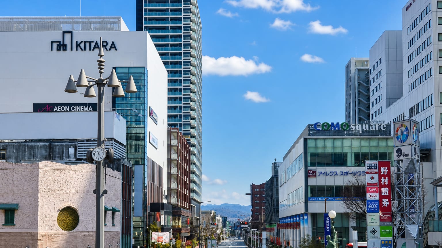 Street view in Toyota, Japan, past the KiTARA complex and COMO Square shopping buildings under a blue sky.