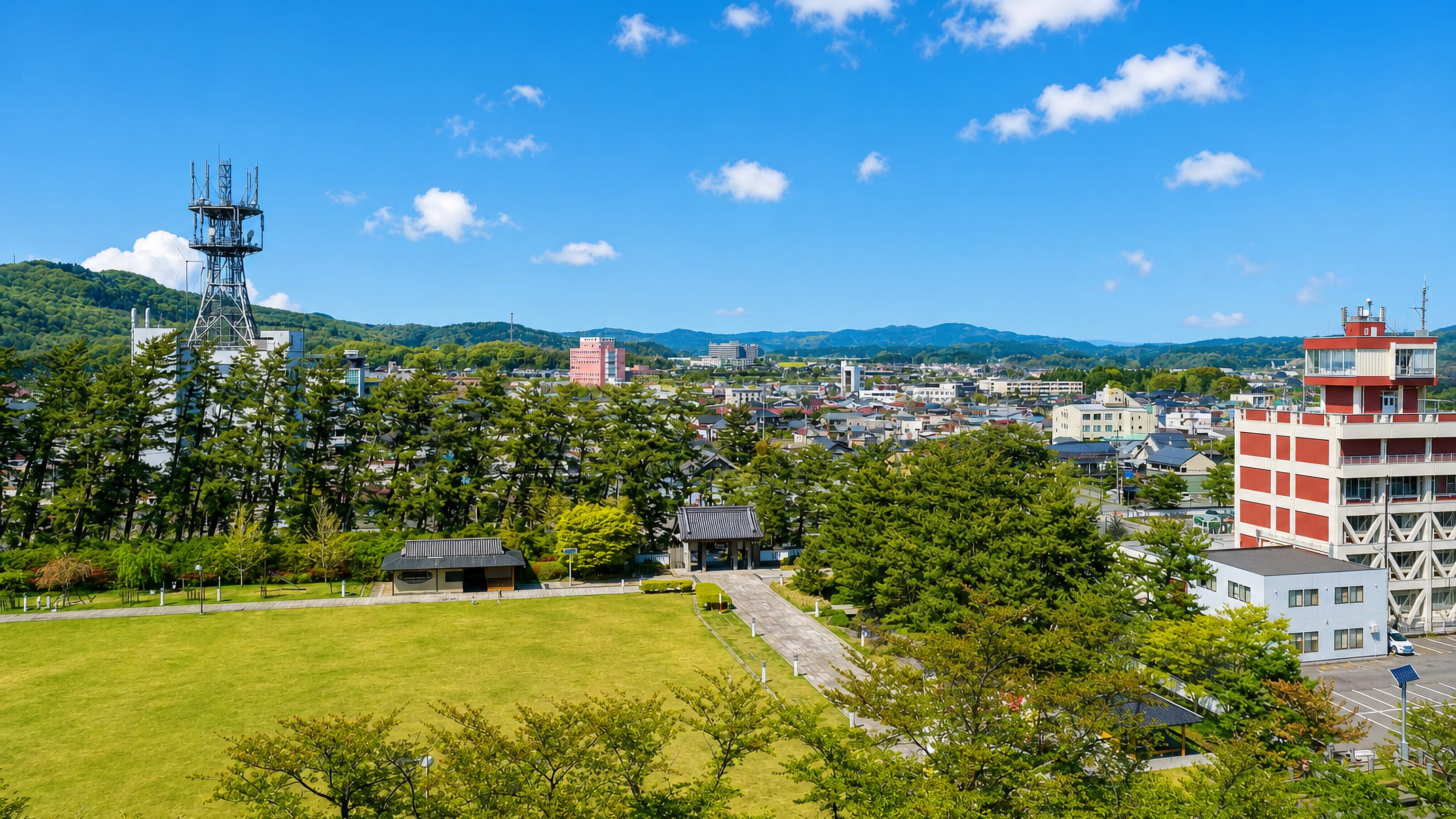 Yurihonjō's low-rise townscape in Akita, seen across a park lawn toward forested hills under a blue sky.