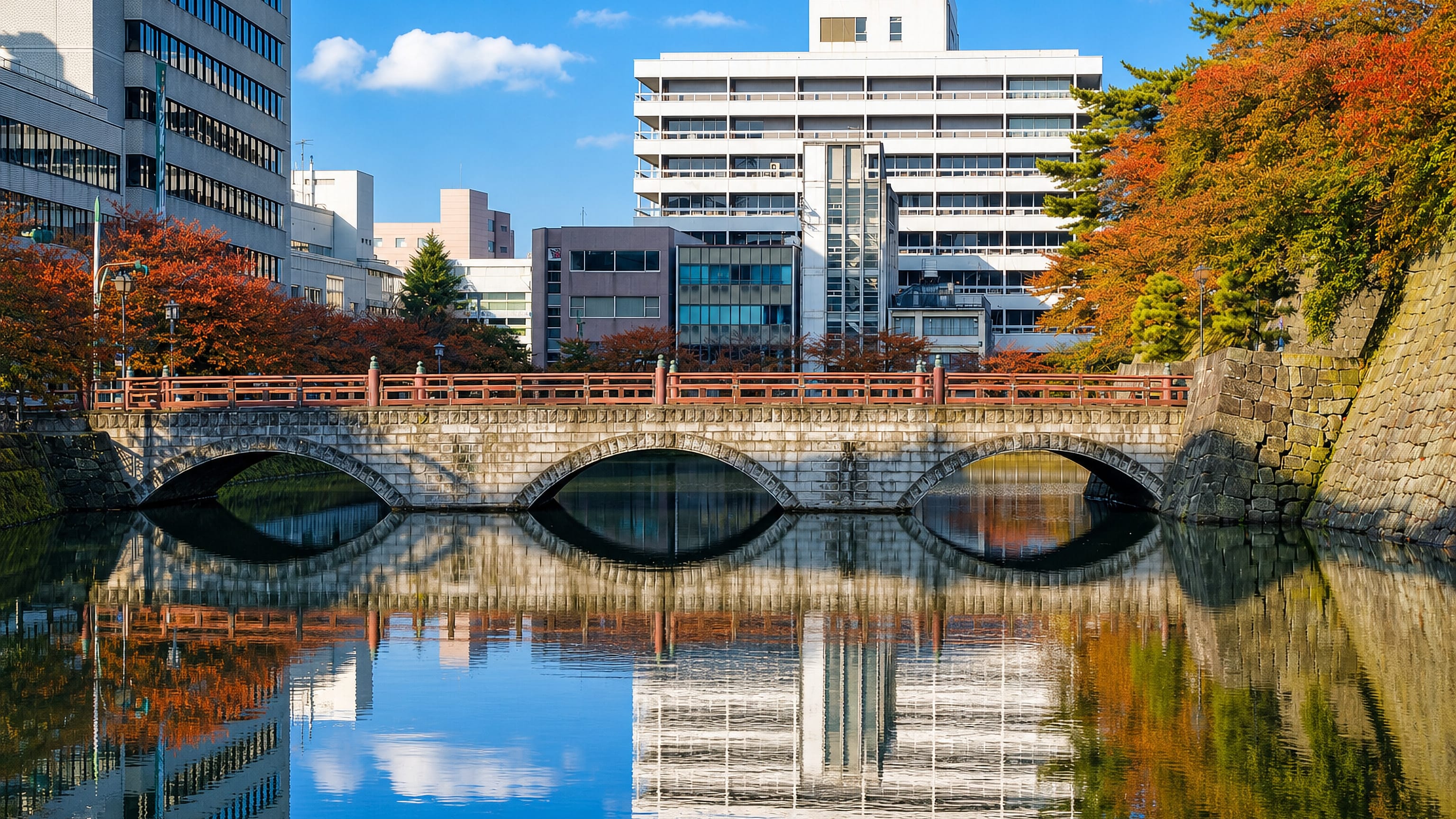 Stone arch bridge with red railings over the Fukui Castle moat, backed by downtown Fukui in autumn.
