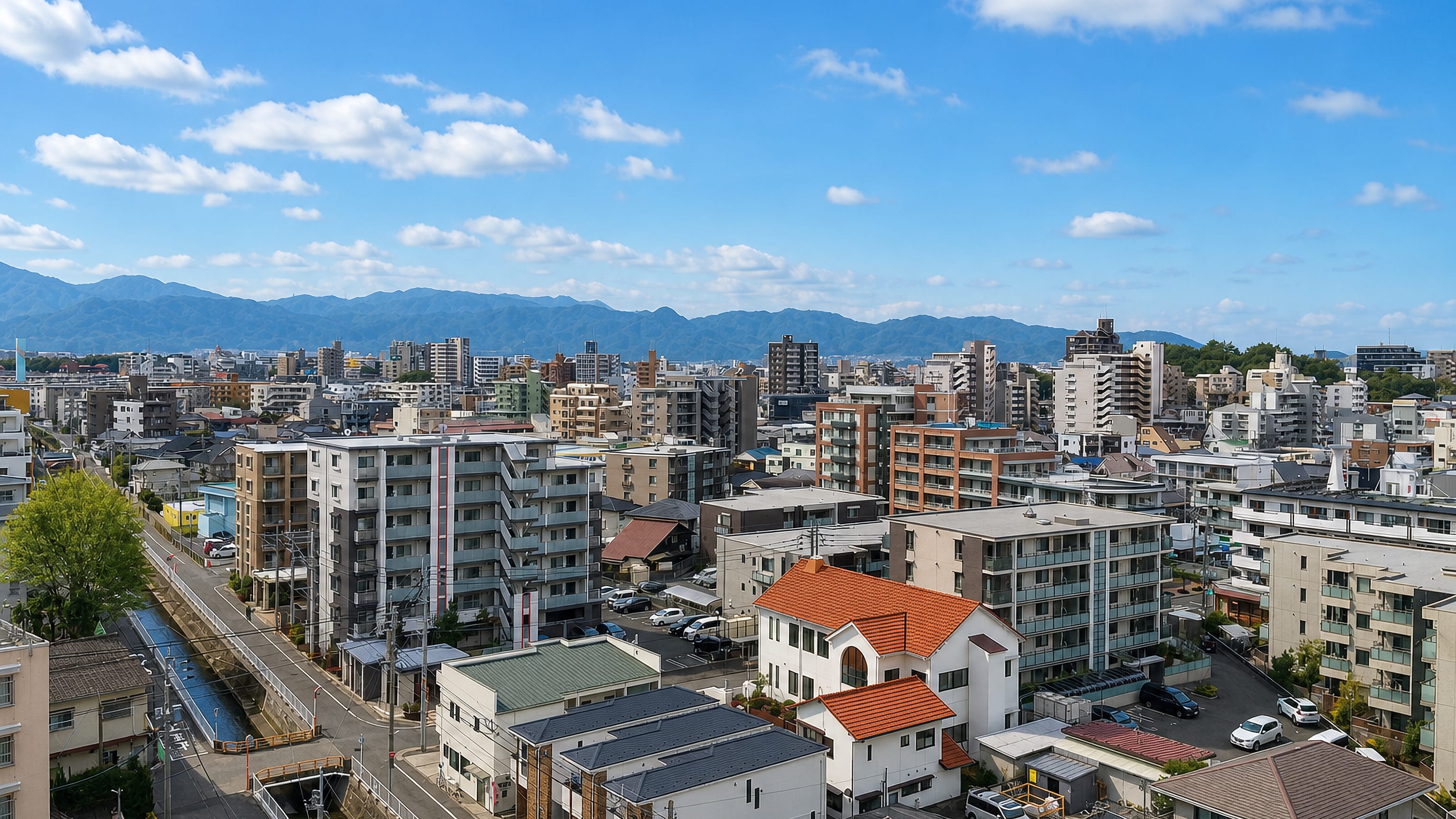 Elevated view over Akebono in Fukuoka: mid-rise apartment blocks, tiled houses and a canal below distant mountains.