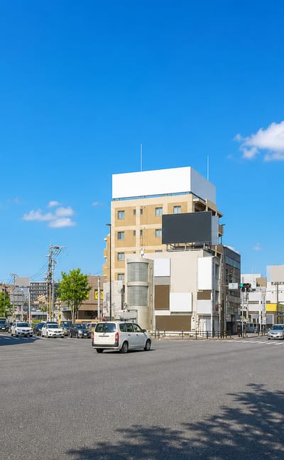 Sunlit crossroads in Hara, Fukuoka, lined with low mid-rise offices and shops under a clear sky.