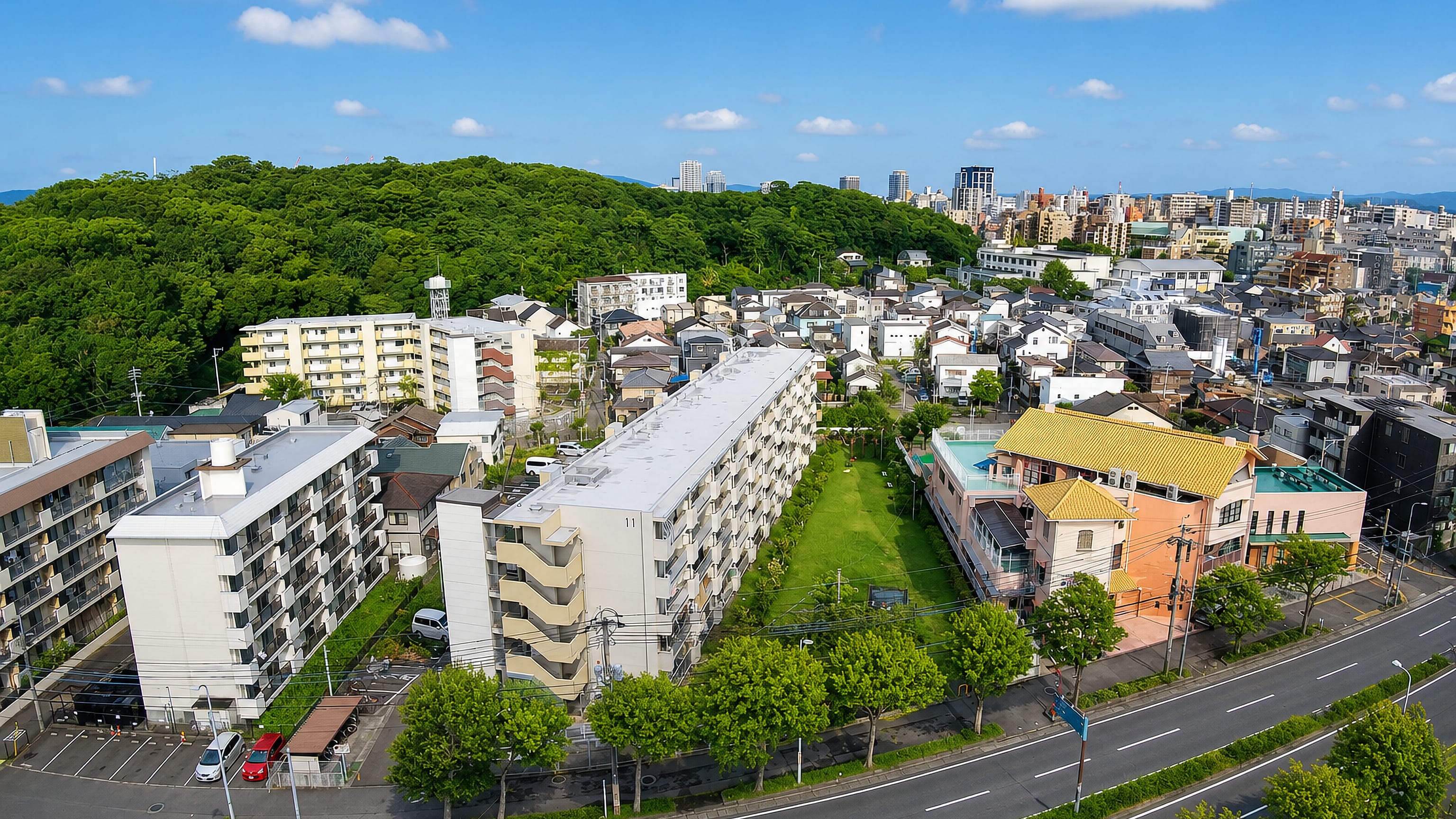 Aerial view of apartment blocks and houses below a wooded hill in Izaki, Fukuoka, with the city skyline beyond.