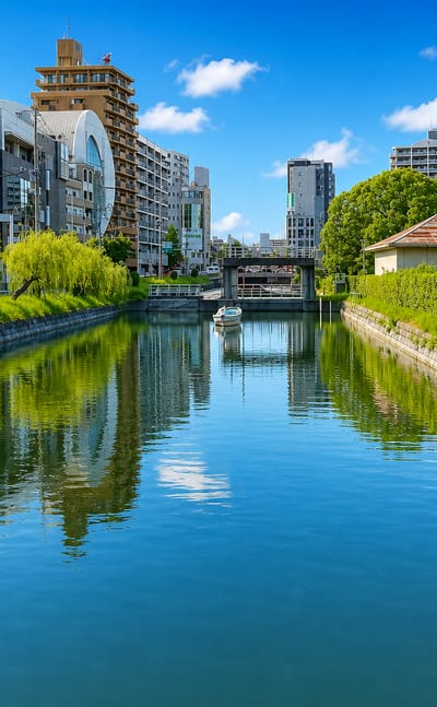 Willow-lined canal reflecting apartment towers and low tiled houses in Kuromon, Fukuoka.