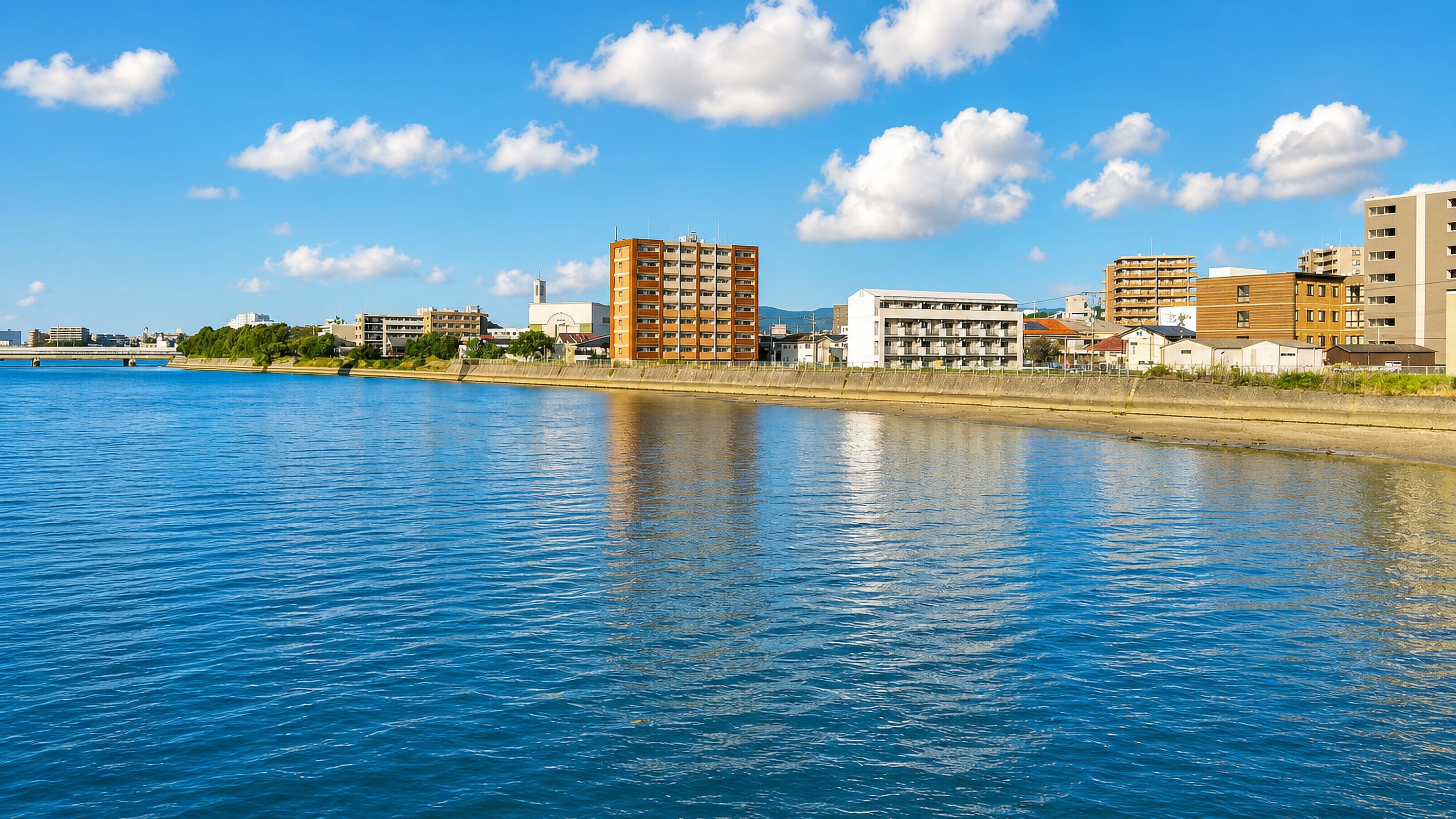 Riverside apartment blocks and seawall at Matsushima in Fukuoka, seen across calm blue water.