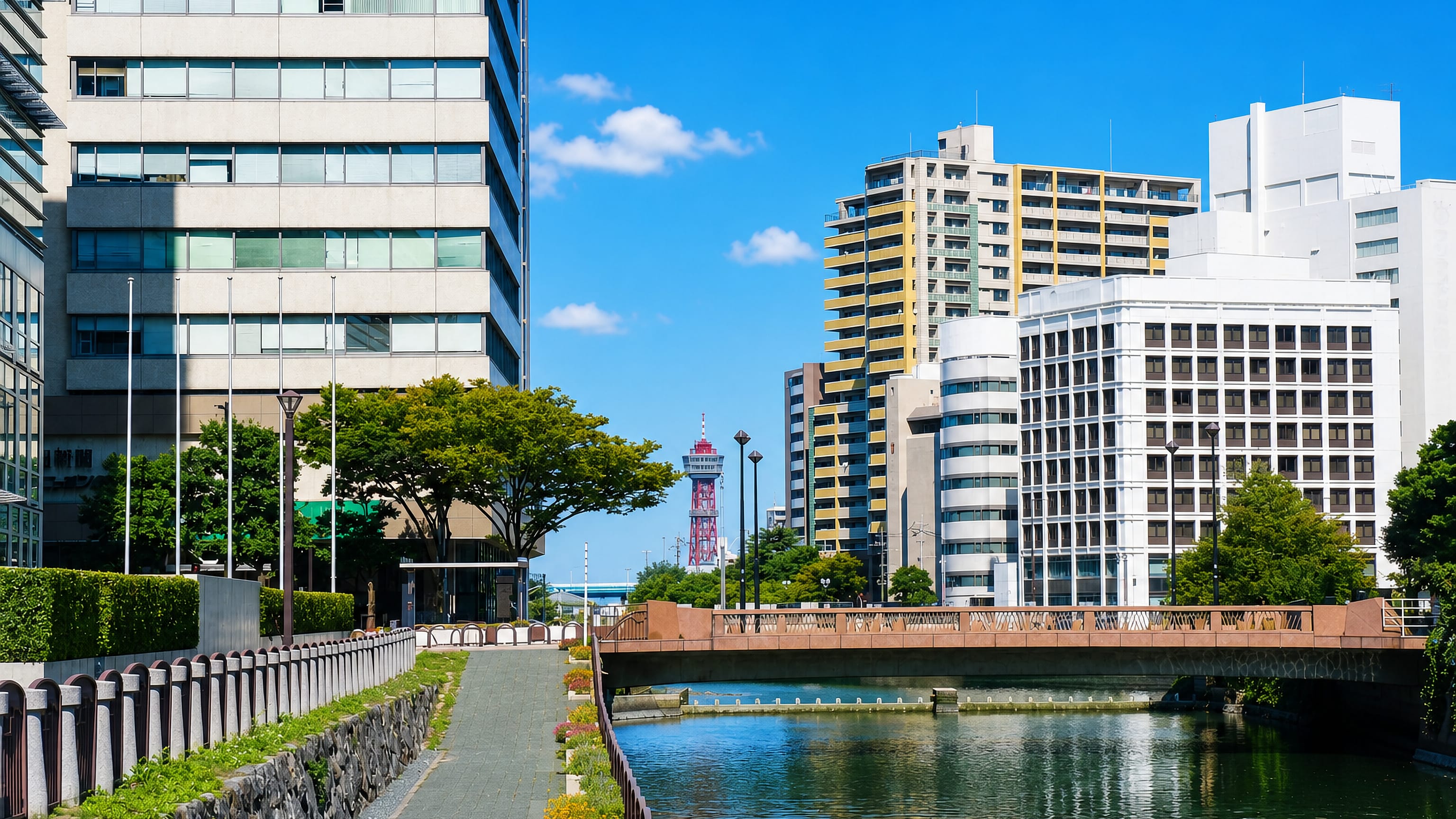 Sunlit canal in Tenjin, Fukuoka, with modern office towers and a red-and-white port tower beyond a stone bridge.