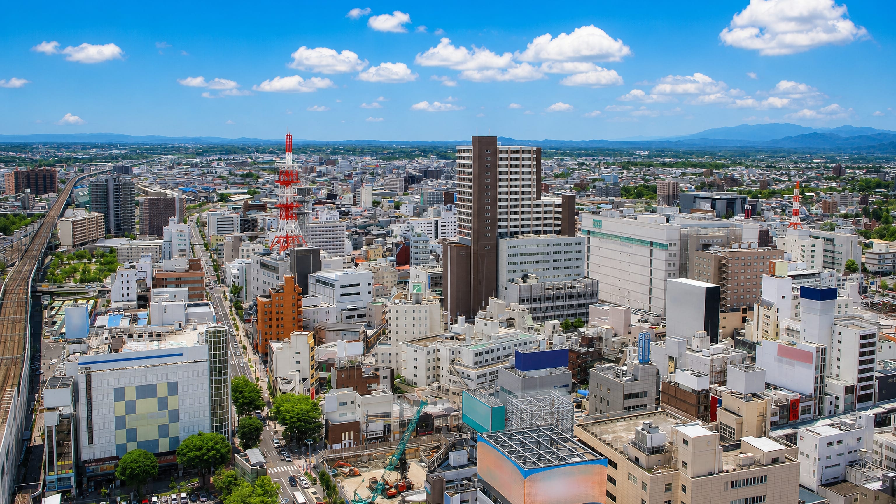 Aerial view over Kōriyama's downtown blocks, red-and-white radio tower and rail lines, with hills beyond.