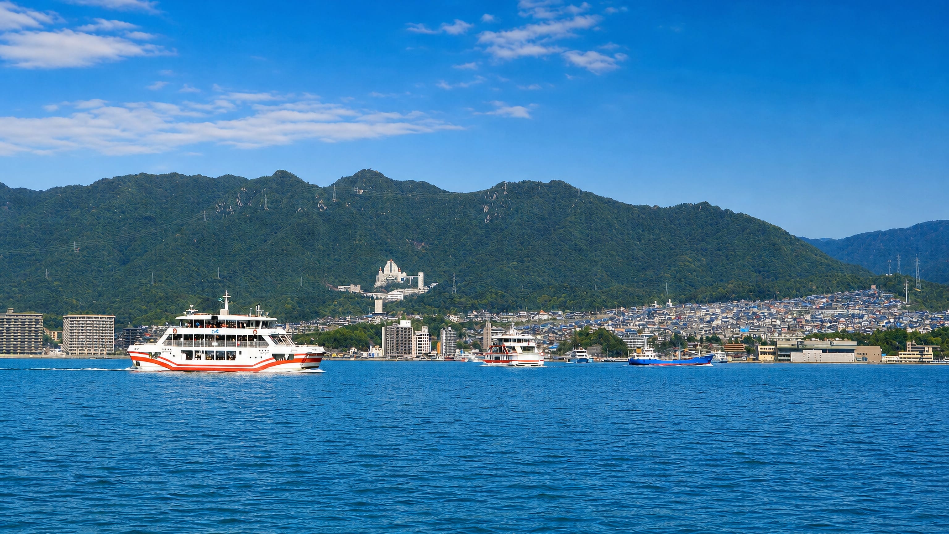 Red-and-white Miyajima ferry crossing the Seto Inland Sea toward Hatsukaichi's hill-backed waterfront.