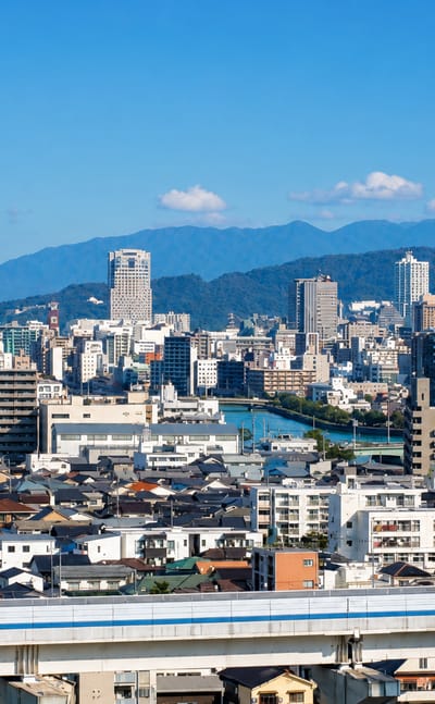 Wide daytime view over Hiroshima's low-rise cityscape and river with a shinkansen train crossing the foreground