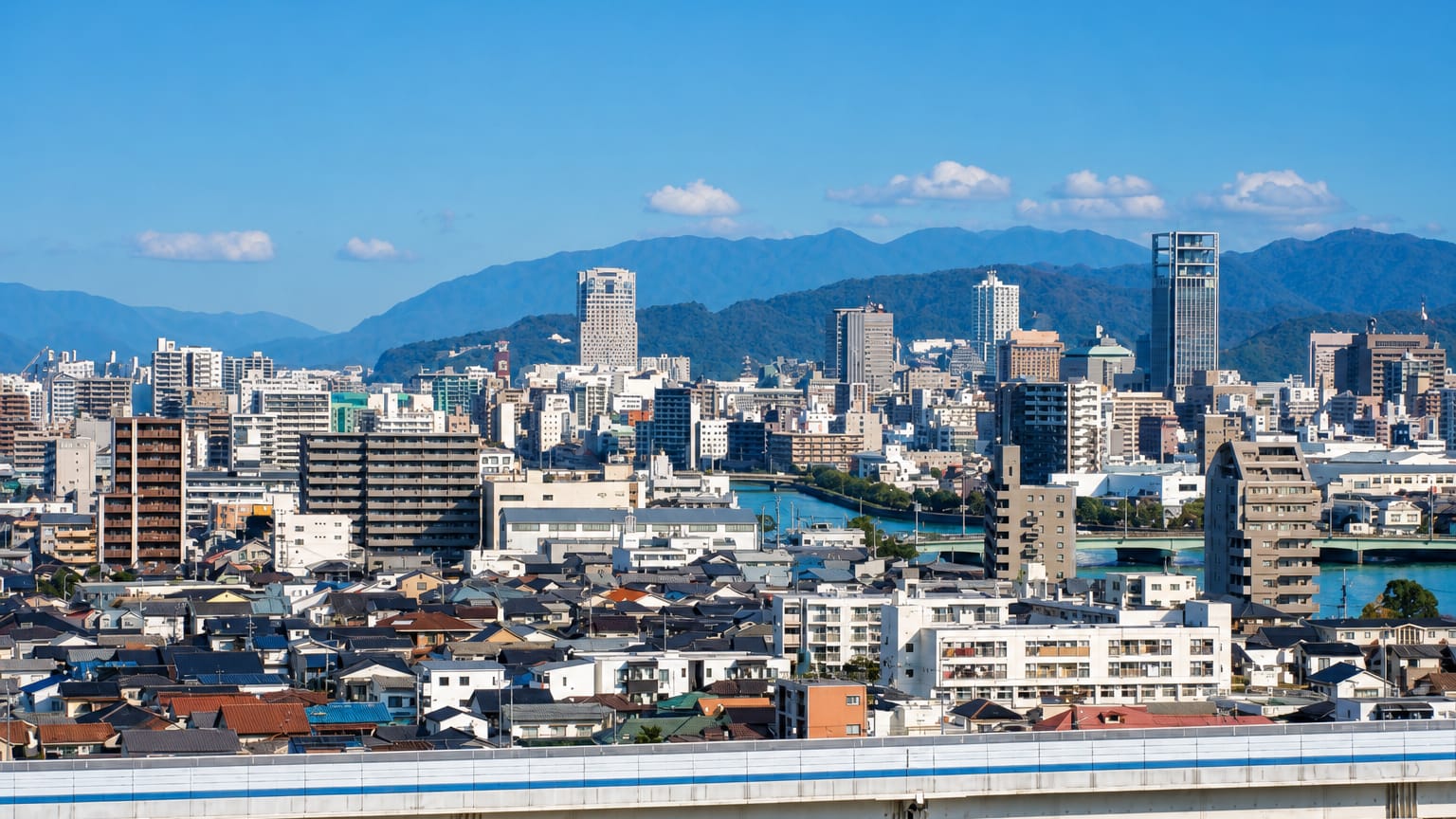 Wide daytime view over Hiroshima's low-rise cityscape and river with a shinkansen train crossing the foreground