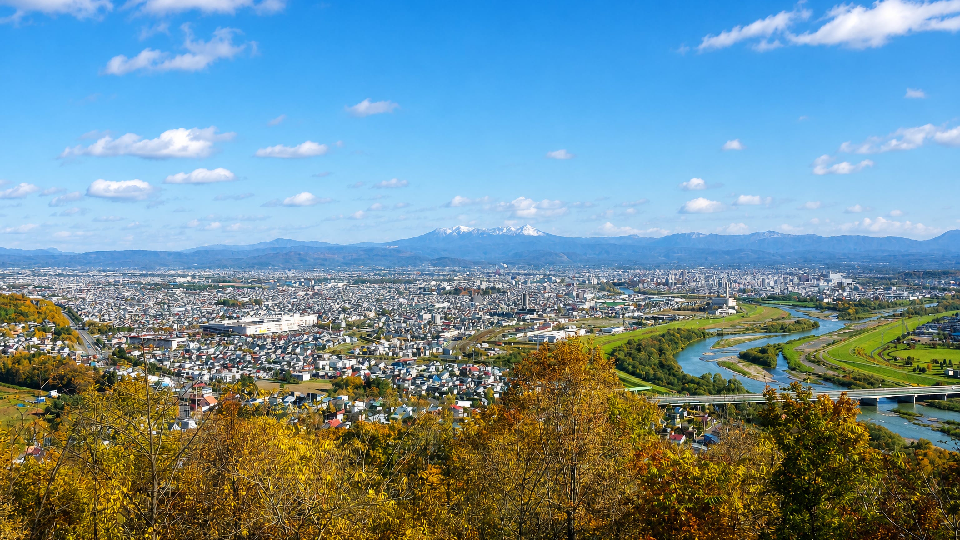 Asahikawa spreading across its river basin in autumn, with the snow-capped Daisetsuzan peaks on the horizon.