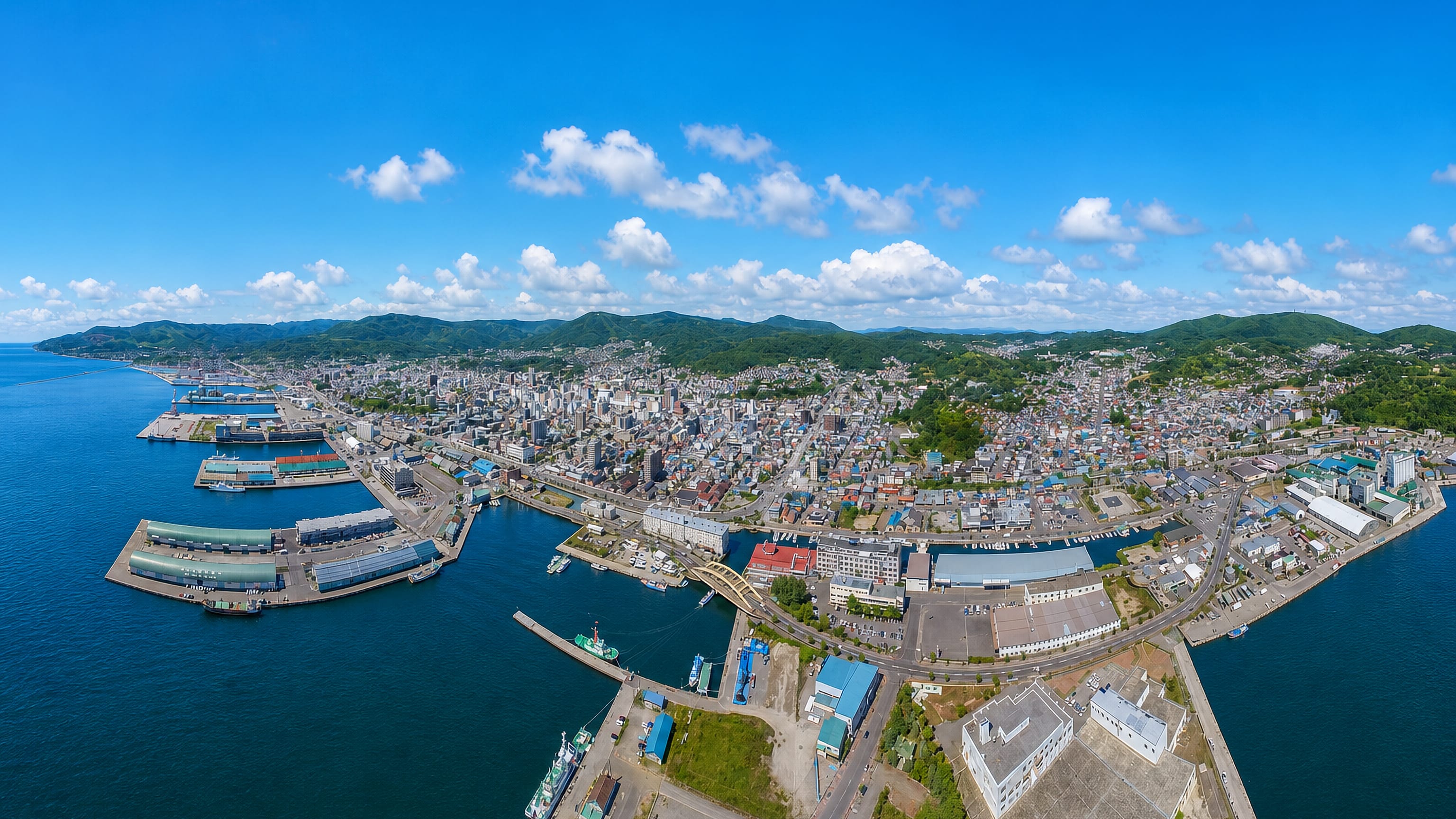 Aerial view of Otaru, Hokkaido, its harbour piers and downtown grid backed by green coastal hills.
