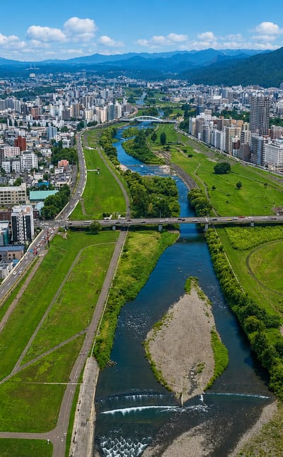 Sapporo's grid of buildings along the Toyohira river with grassy banks and mountains rising behind