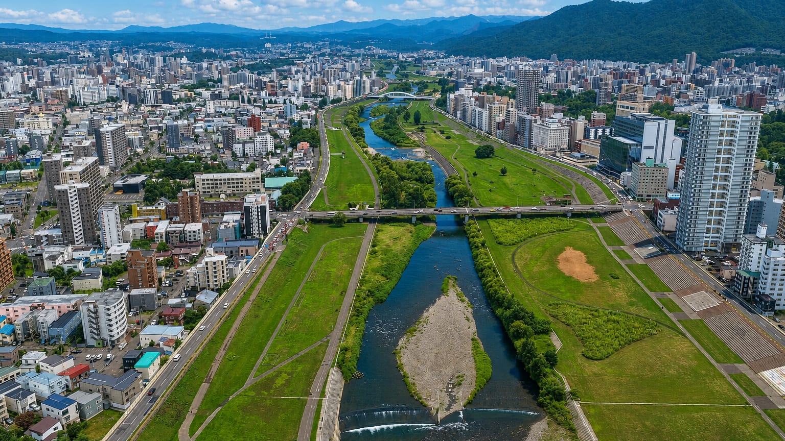 Sapporo's grid of buildings along the Toyohira river with grassy banks and mountains rising behind