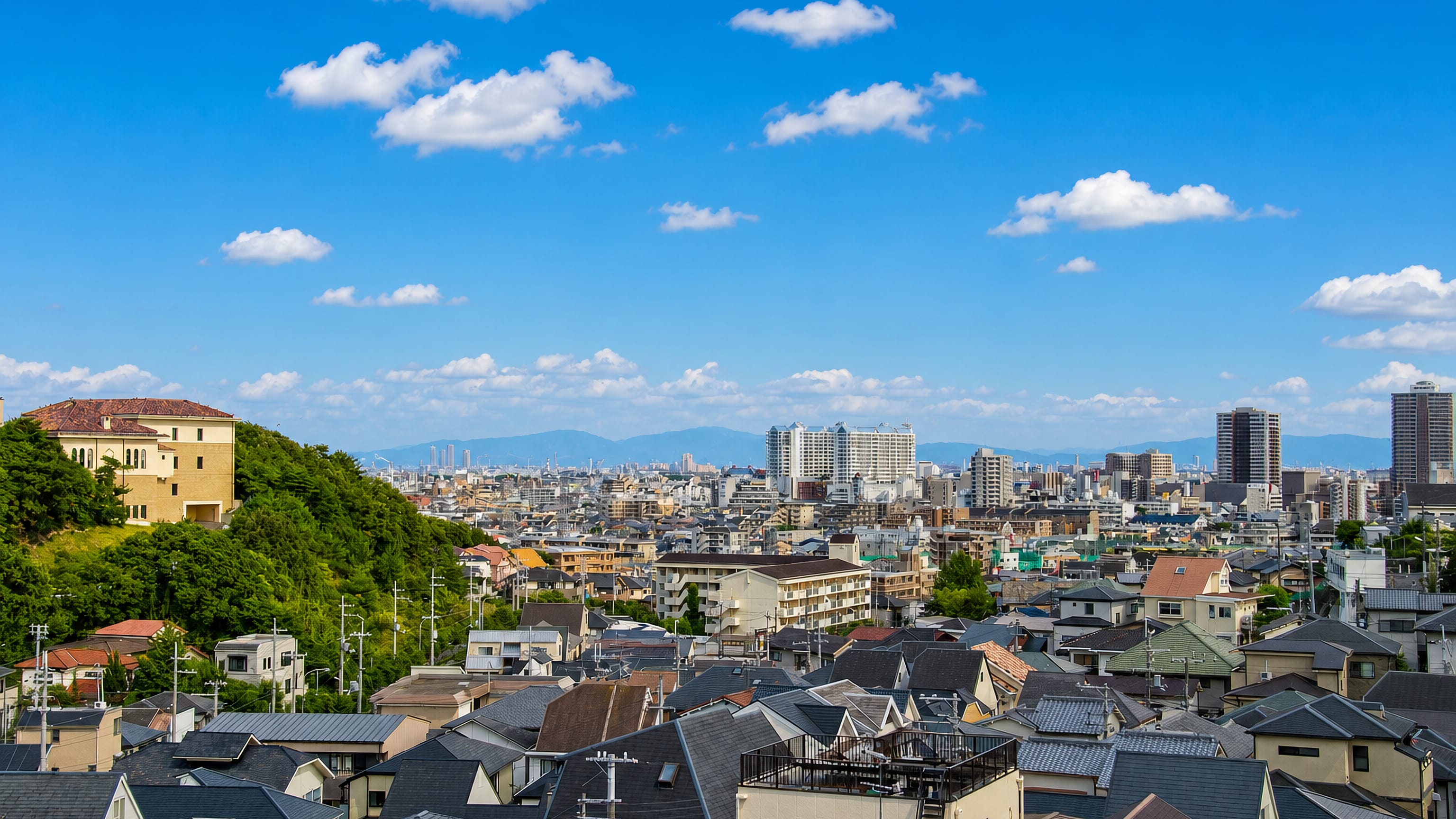 Nishinomiya, Japan, with tiled residential rooftops, a wooded hilltop building and distant high-rises below mountains.