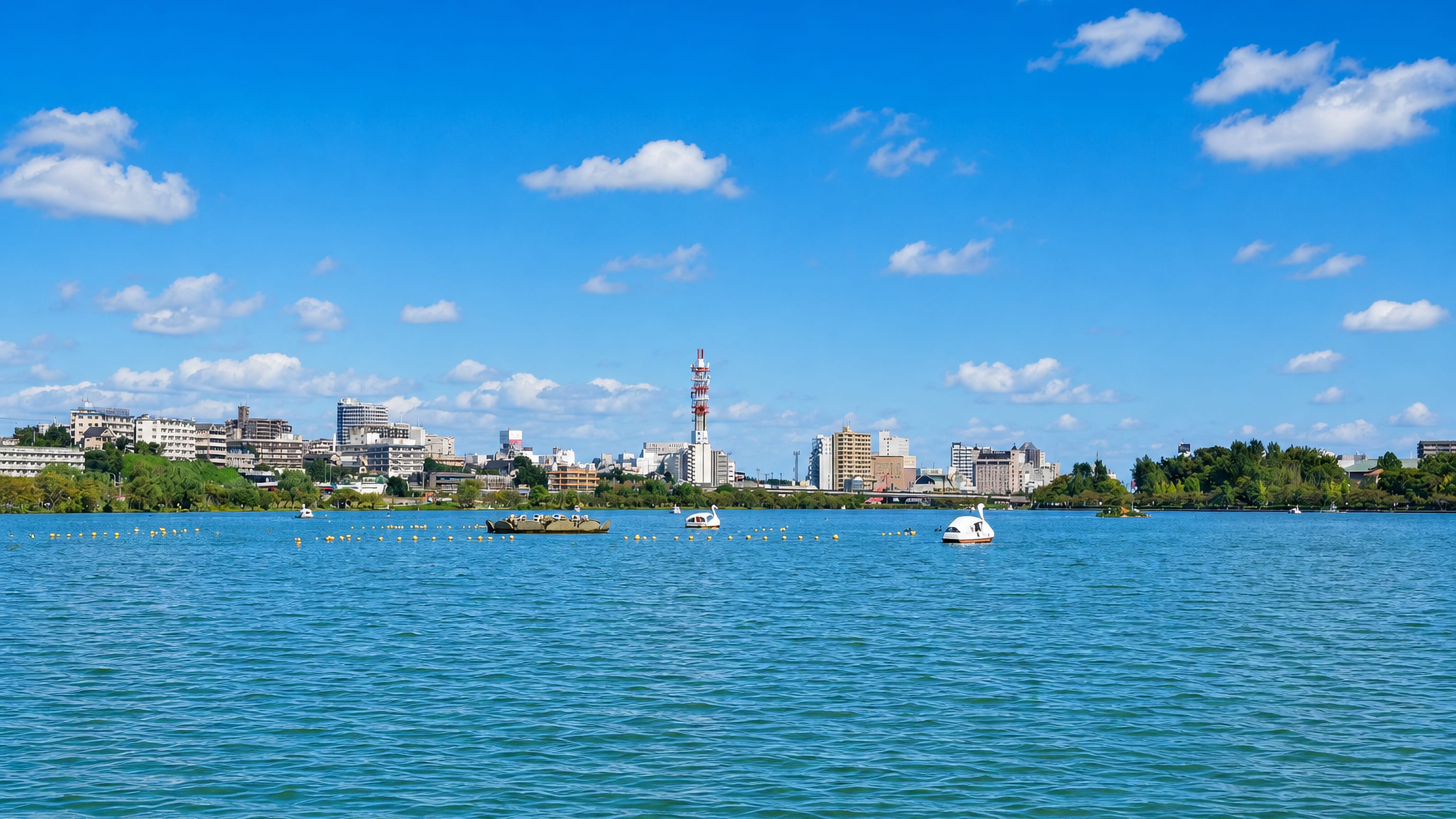 Mito's low skyline and TV mast across Lake Senba, with swan pedal boats on the water under a clear sky.