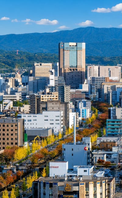 Kanazawa cityscape in autumn, a tree-lined boulevard running through mid-rise buildings toward forested mountains.