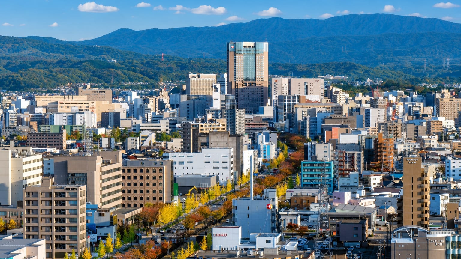 Kanazawa cityscape in autumn, a tree-lined boulevard running through mid-rise buildings toward forested mountains.