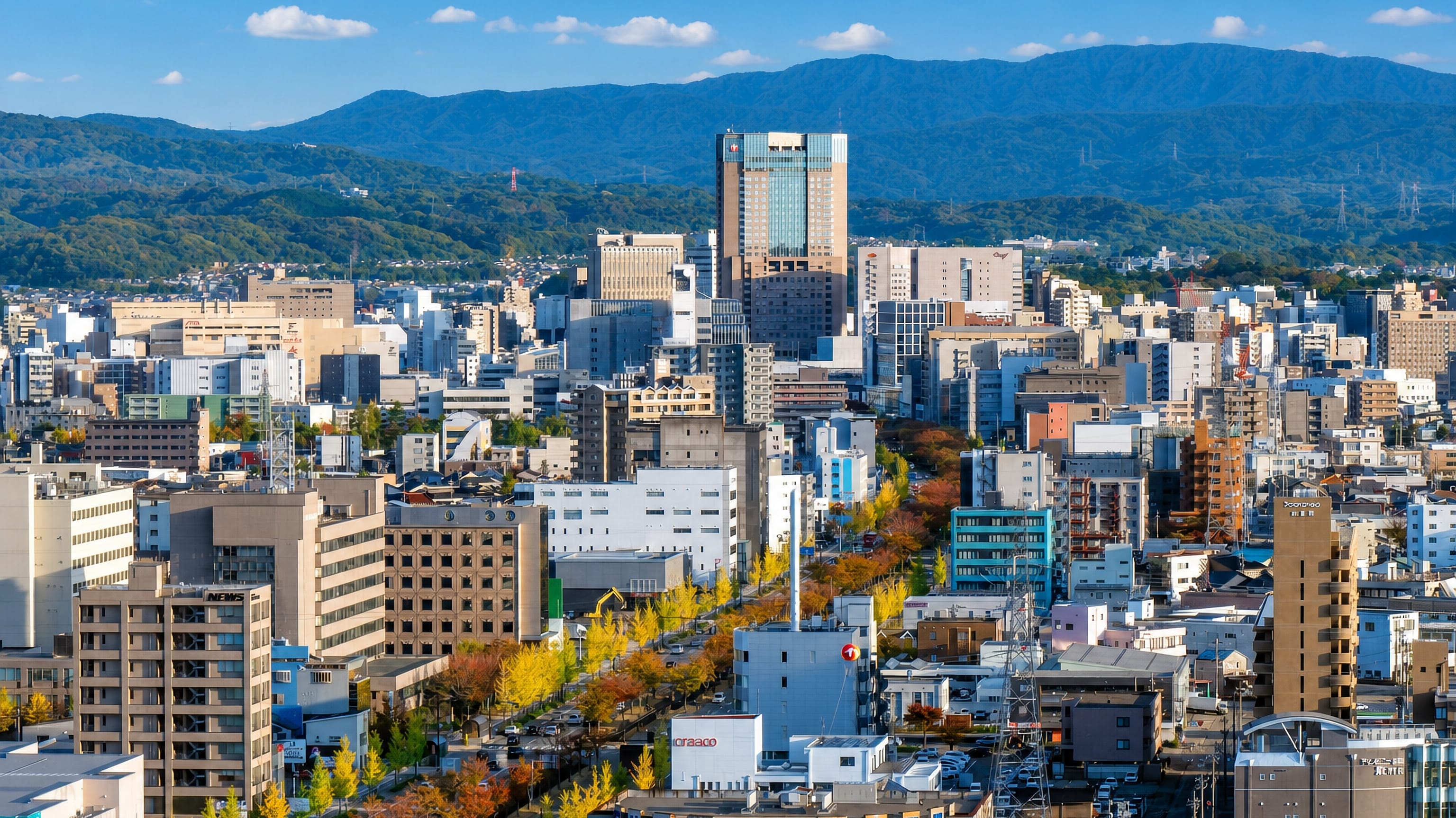 Kanazawa cityscape in autumn, a tree-lined boulevard running through mid-rise buildings toward forested mountains.