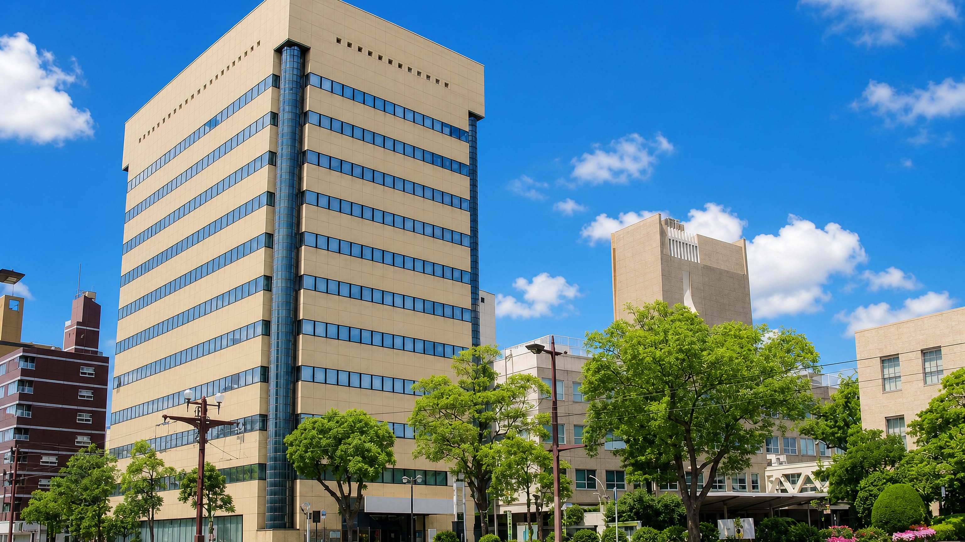A modern office tower and tree-lined avenue in Yamashitacho, central Kagoshima, under a clear blue sky.