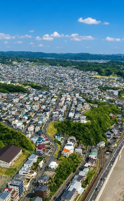 Aerial view of Kamakura's coastline, houses on green hills above a curving sandy beach along the blue sea.