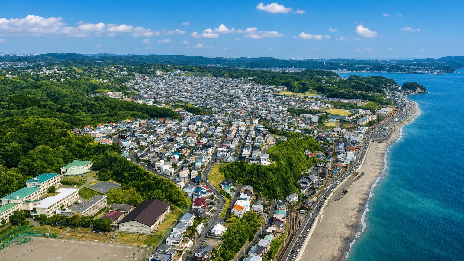 Aerial view of Kamakura's coastline, houses on green hills above a curving sandy beach along the blue sea.