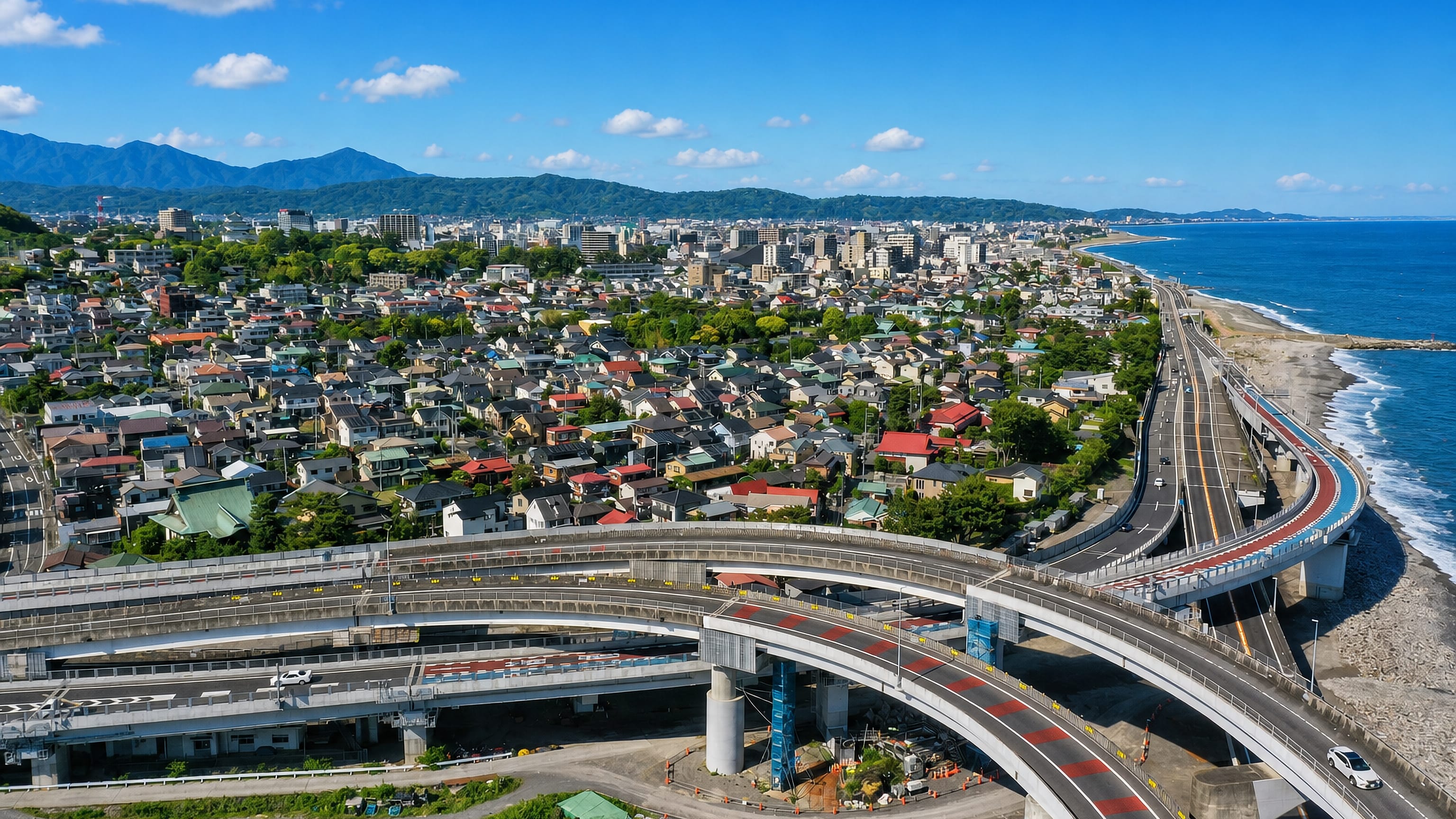 Aerial view of Odawara, Japan, its coastal expressway interchange curving along Sagami Bay below forested hills.