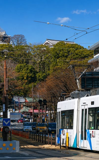Kumamoto Castle on its wooded hill above a city street, with a Kumamoto tram bound for Kengunmachi.
