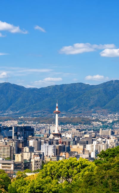 Kyoto cityscape with the red-and-white Kyoto Tower rising above the buildings against forested mountains.