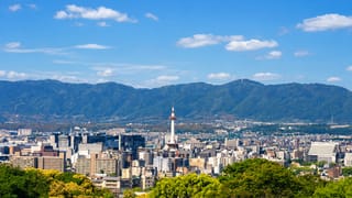 Kyoto cityscape with the red-and-white Kyoto Tower rising above the buildings against forested mountains.