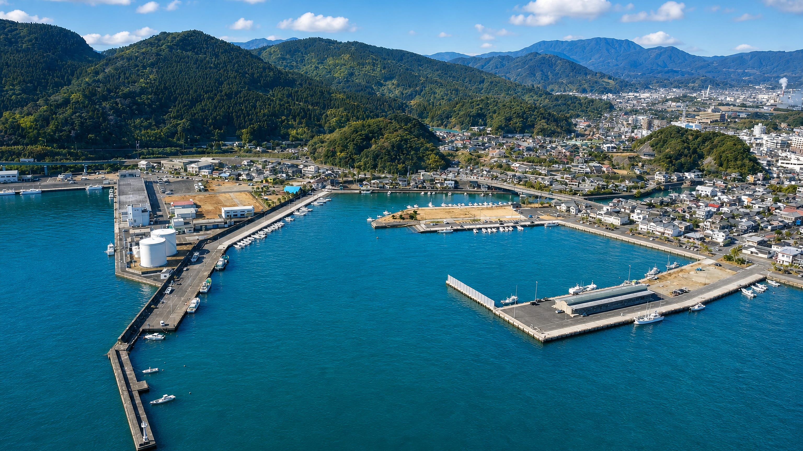 Aerial view of Nichinan's harbour breakwaters and fishing fleet below forested hills in Miyazaki, Japan.