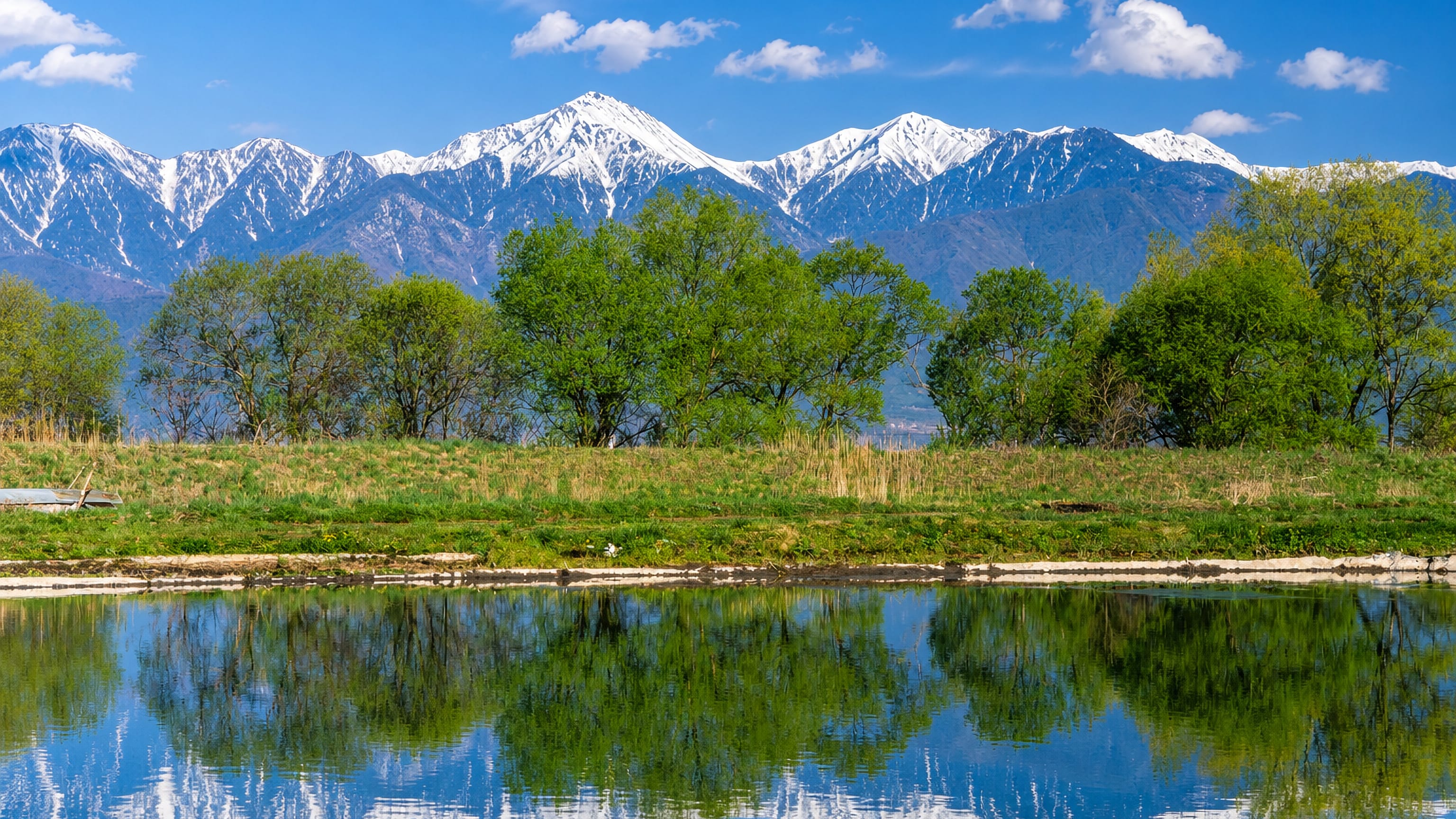 Snow-capped Japanese Alps reflected in a still pond at Azumino, Nagano, framed by fresh spring greenery.