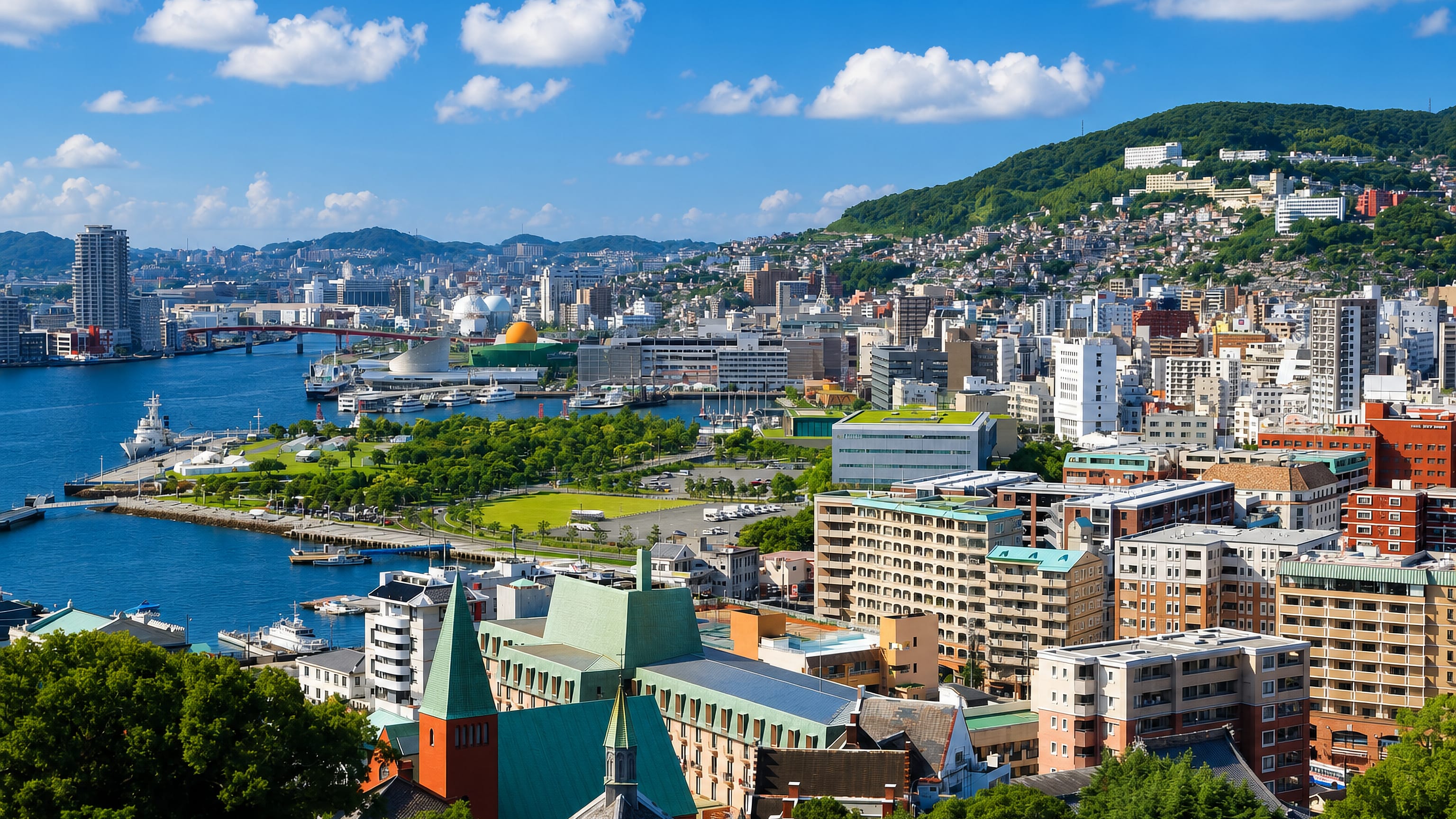 Nagasaki harbour viewed from a wooded hillside, with waterfront parkland and housing climbing the green slopes.