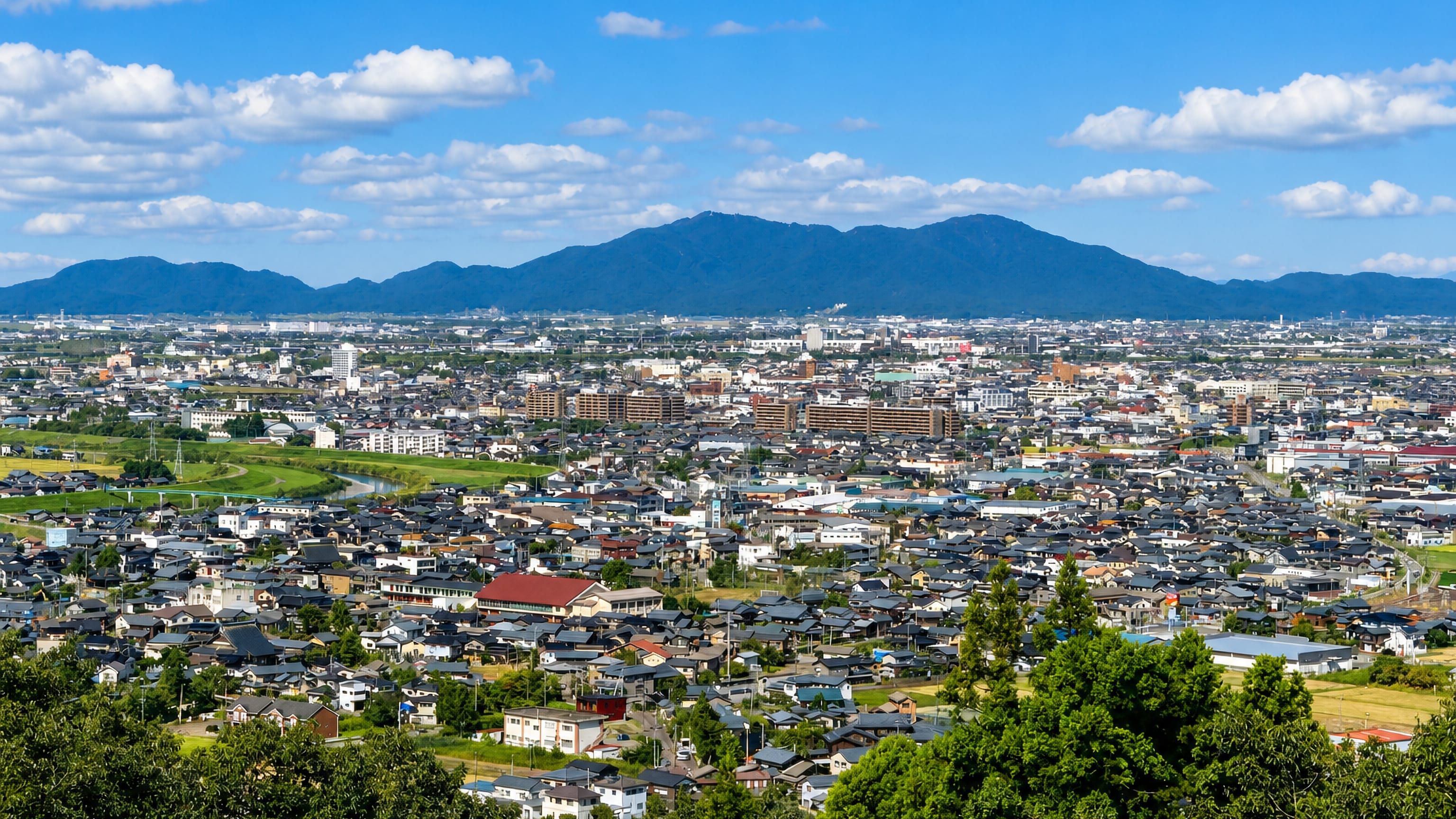 Hillside panorama of Sanjo, Niigata, its dense low-rise townscape spreading to a river and distant mountains.