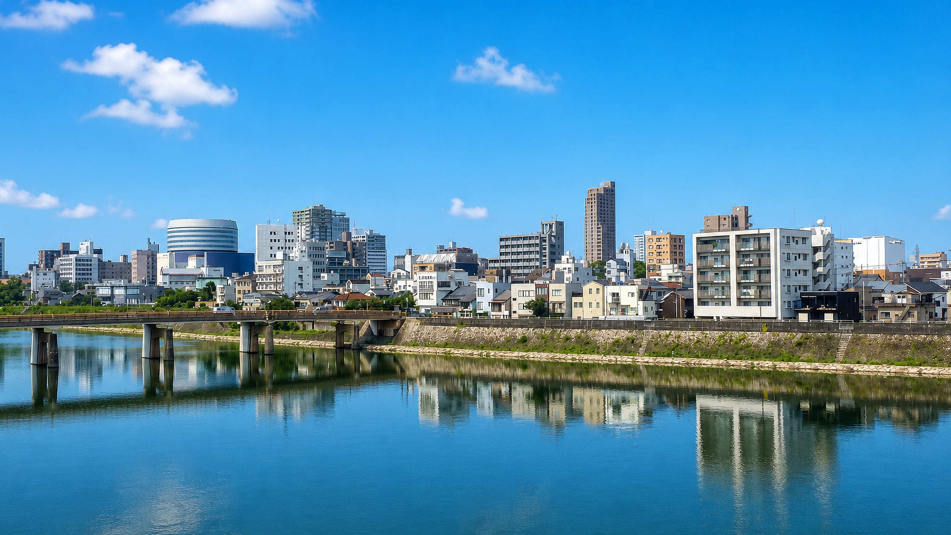 Okayama, Japan, seen across a calm river, with mid-rise blocks, houses and a low road bridge under blue sky