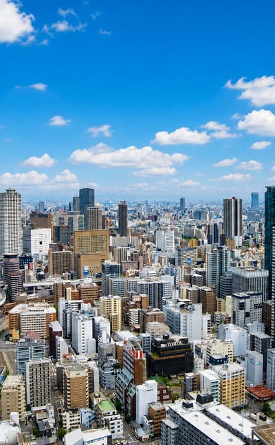 Aerial view over Osaka's dense downtown skyline of high-rise towers and elevated roads under a bright blue sky